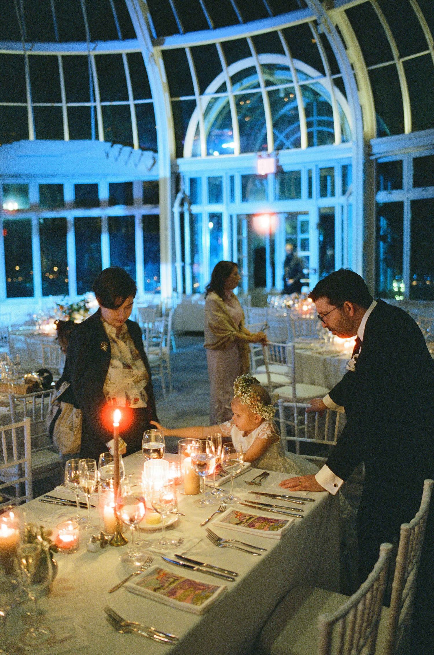 Candlelit dinner setting in an elegant conservatory with arched glass windows and white decor at night.