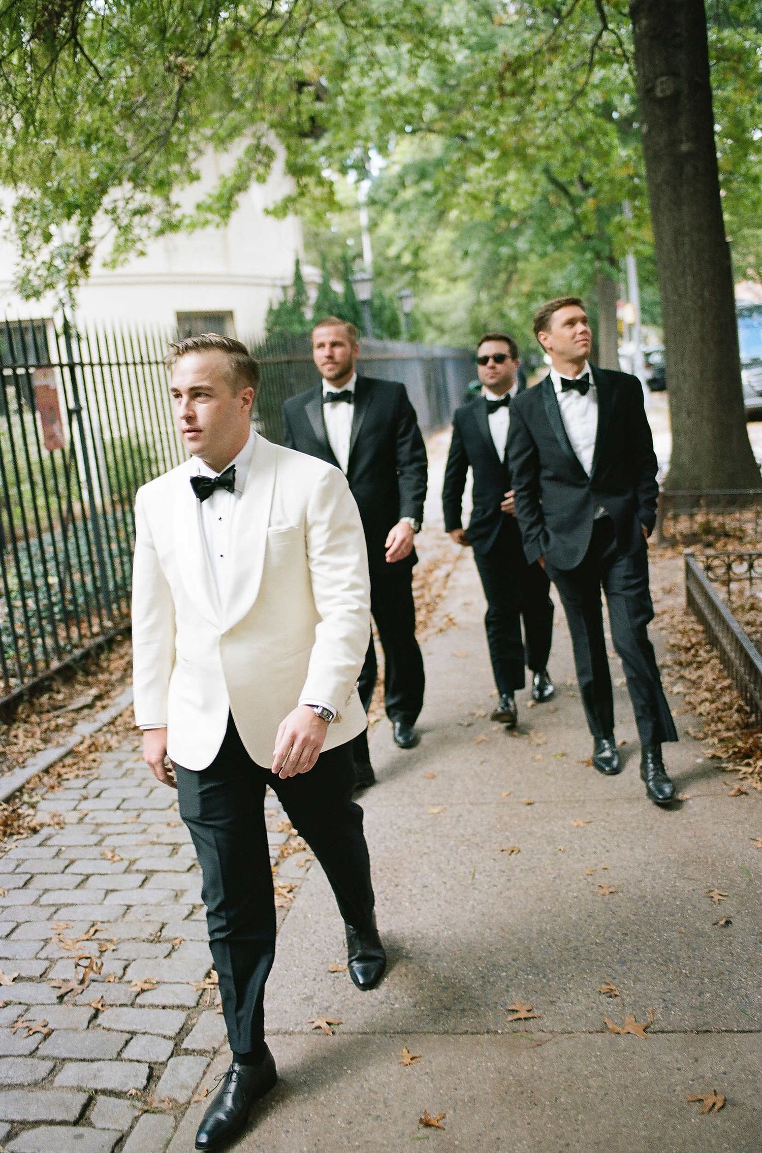 A group in formal black and white tuxedos walk along a tree-lined city sidewalk on an autumn day.