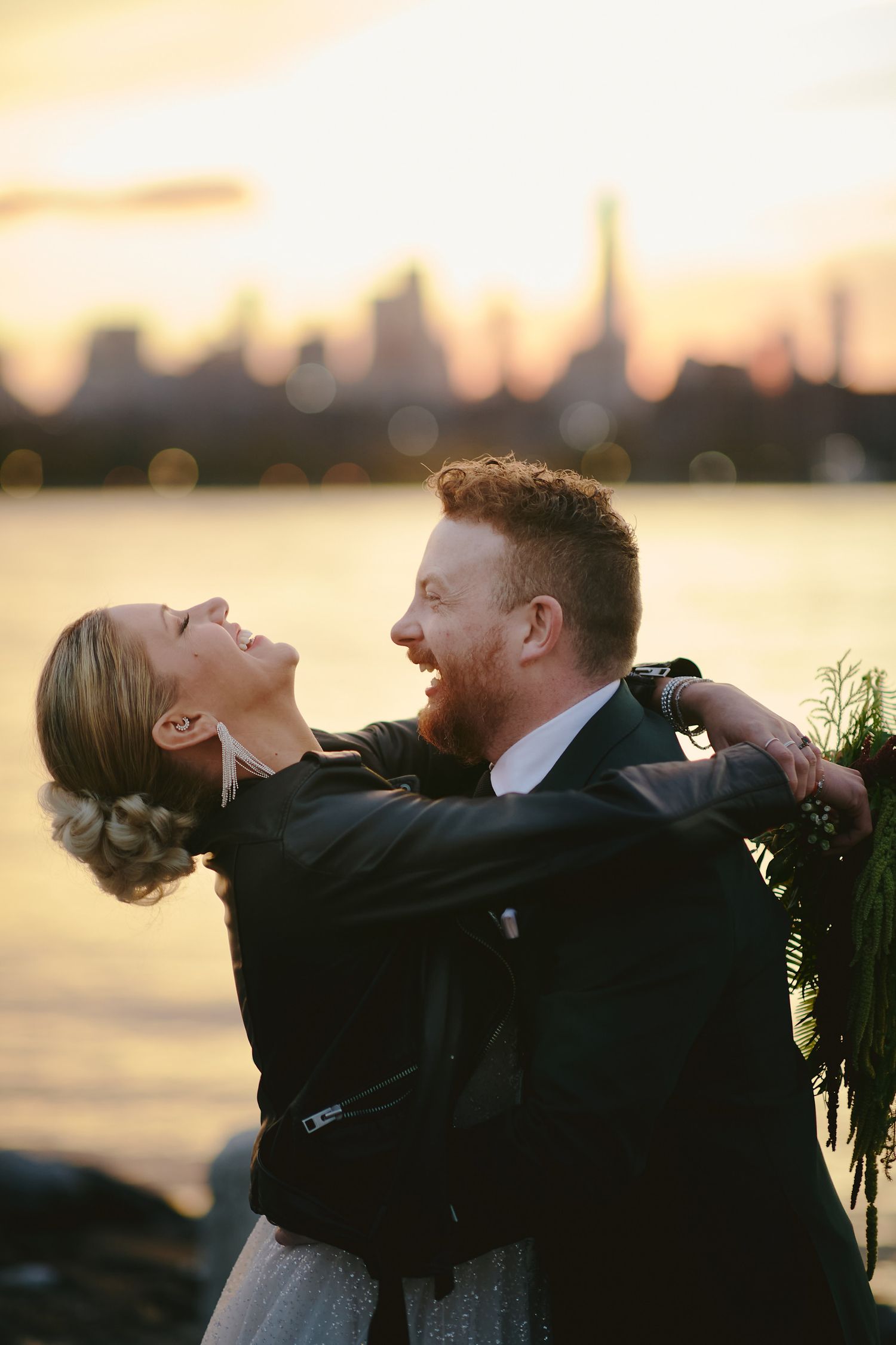A couple embraces and laughs together at sunset with a city skyline silhouette glowing in the background.