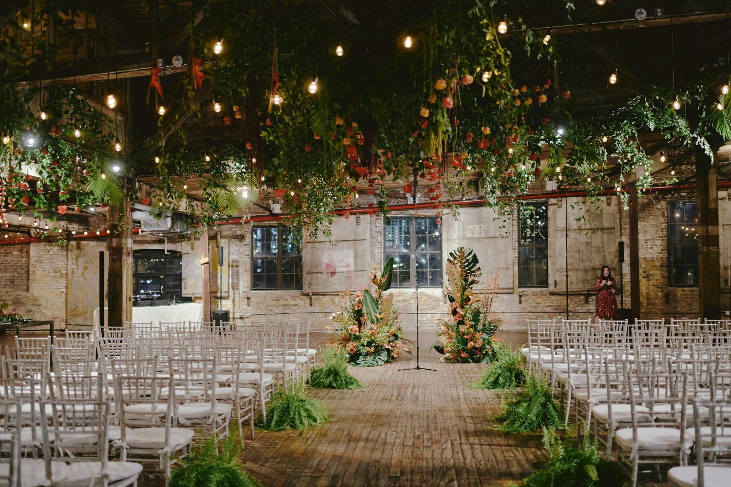 Wedding ceremony setup in a rustic indoor venue with string lights, greenery and white chairs along wooden floors.