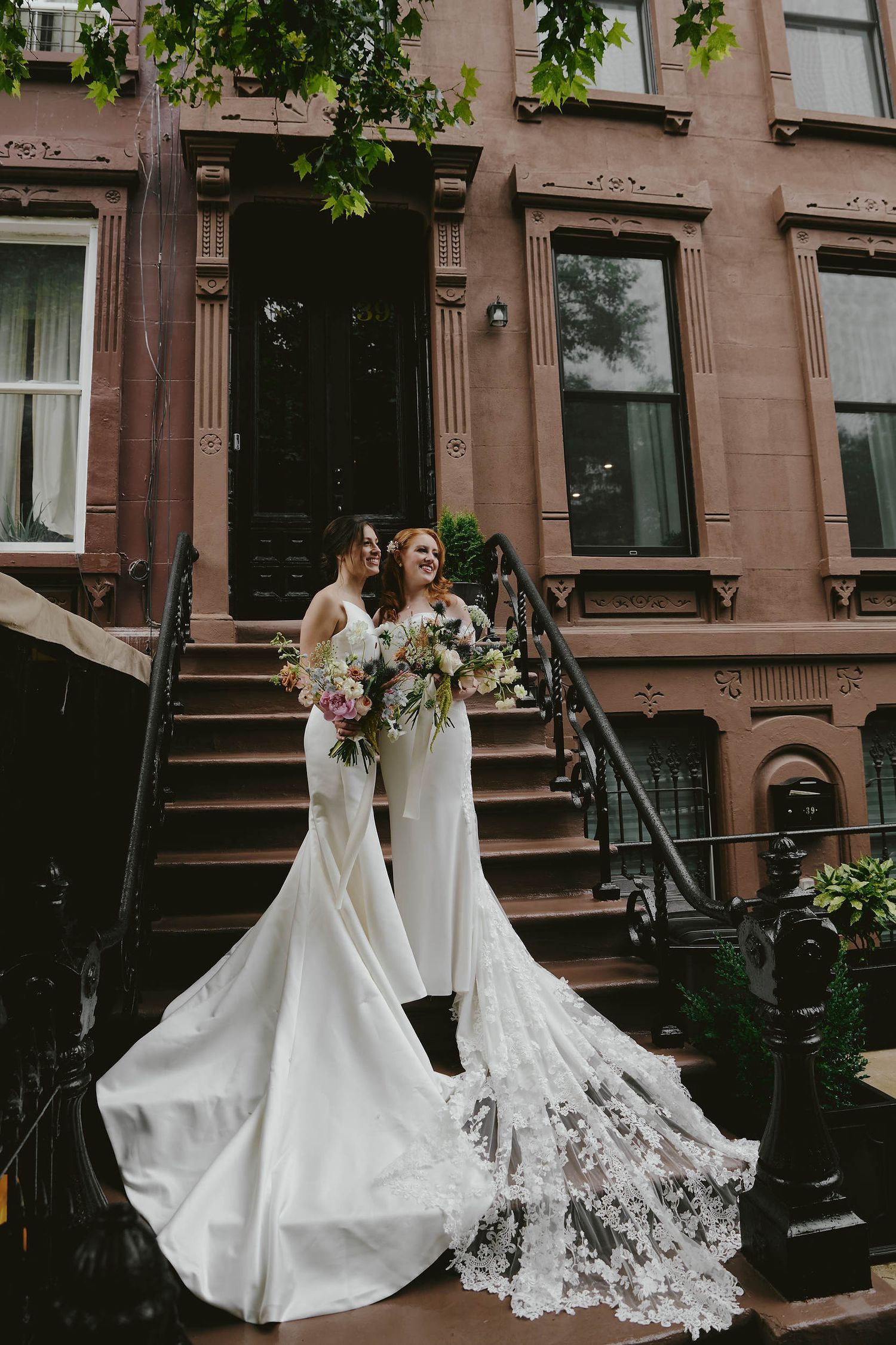 Two people in elegant white wedding gowns with flowing trains stand on brownstone steps in a city neighborhood.