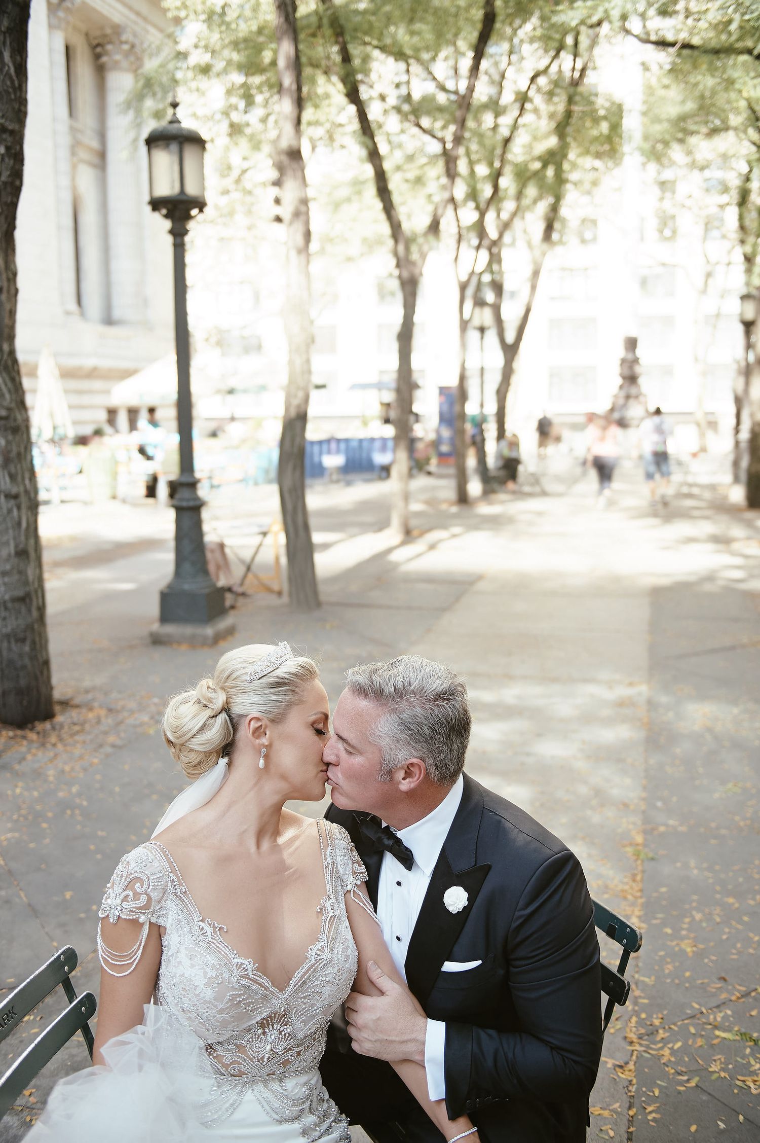 A romantic moment shared between a couple in formal wedding attire on a park bench with lamp posts and trees in the background.