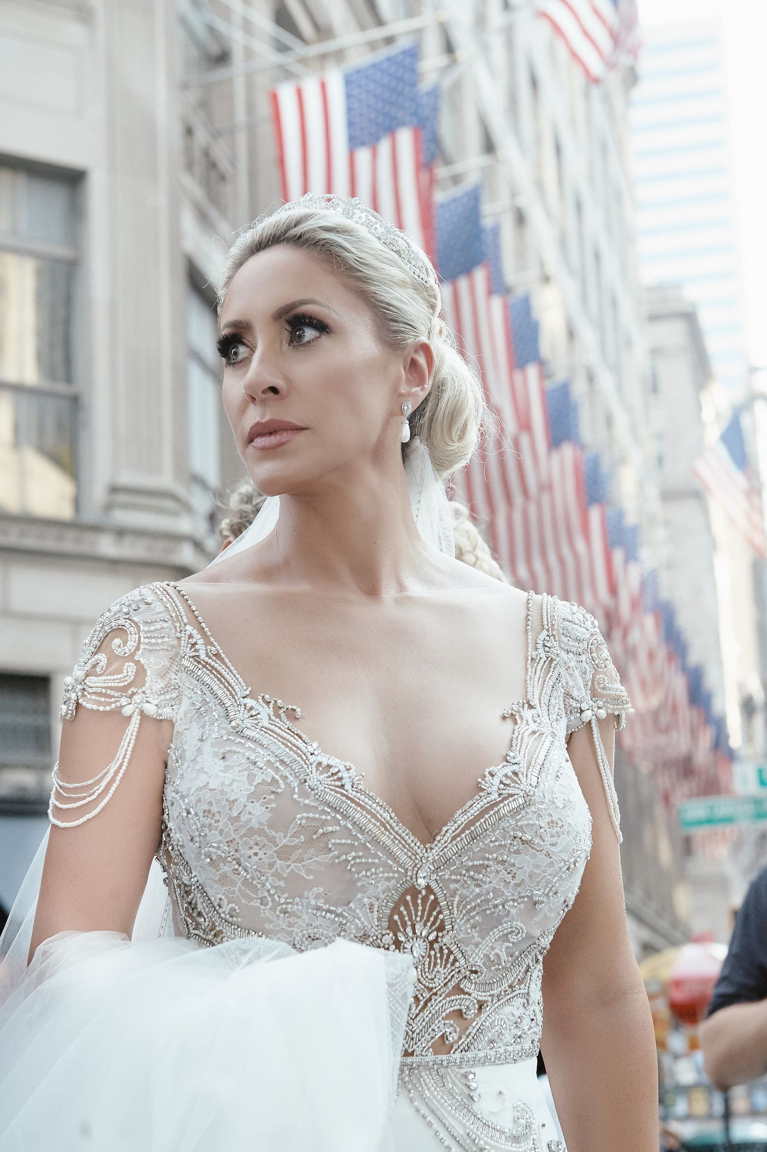 Ornate white lace wedding gown with beaded cap sleeves and American flags visible in the urban background.