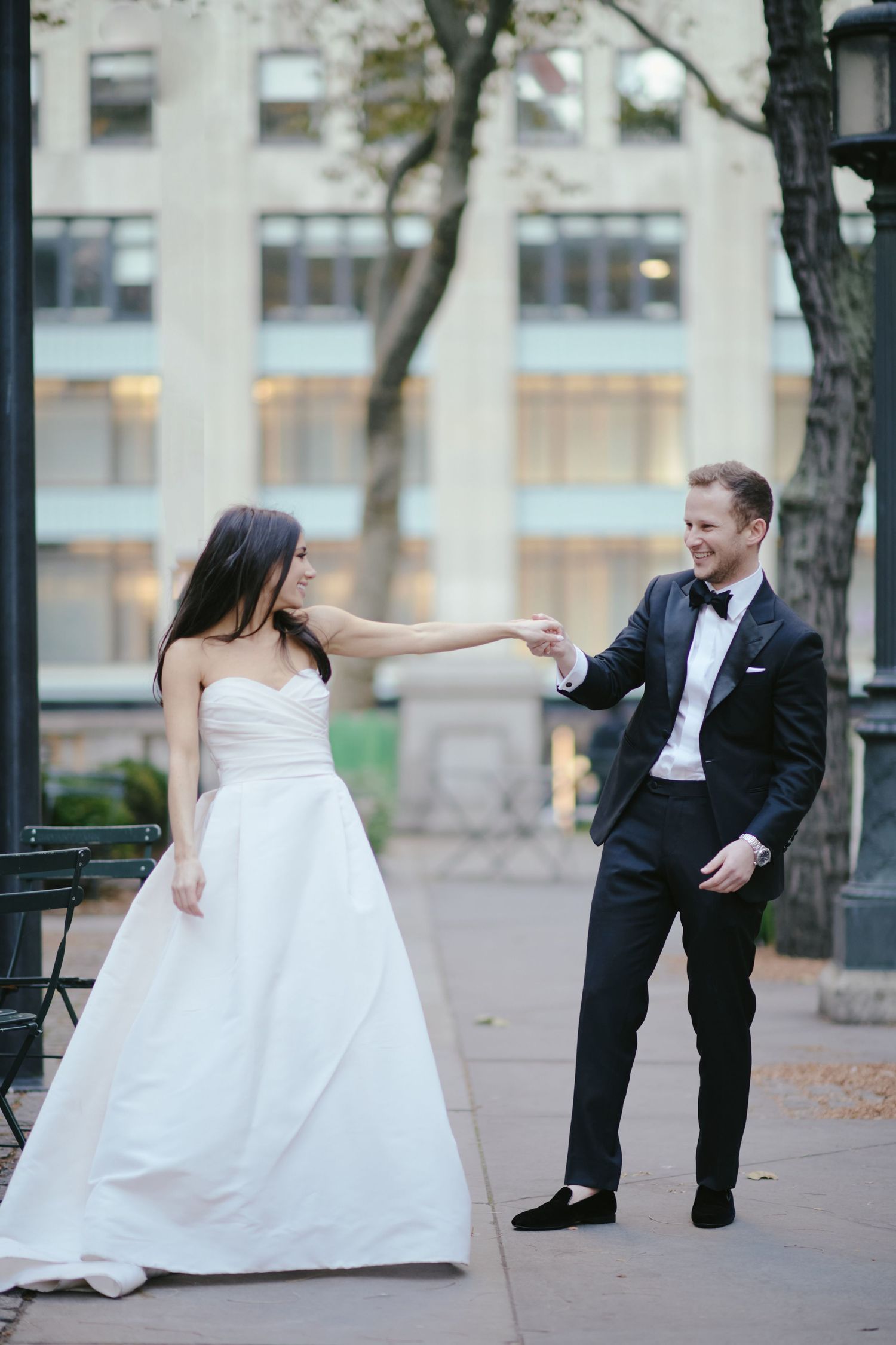 A couple in wedding attire playfully interact on a city sidewalk with trees and buildings in the background.