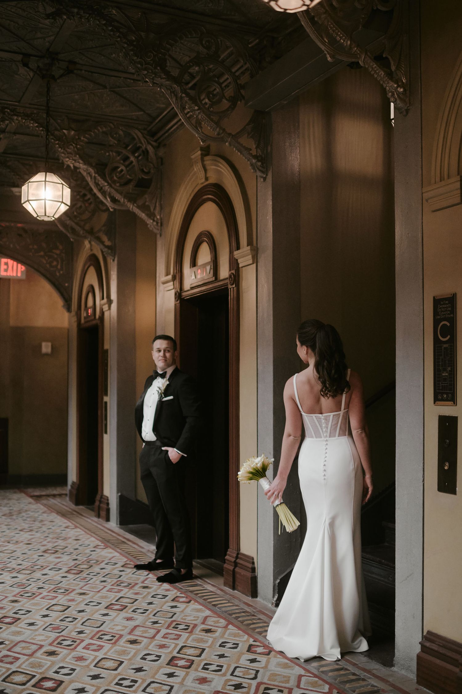A bride in a fitted white dress and groom in a tuxedo stand separated in an ornate vintage hallway with globe lights.