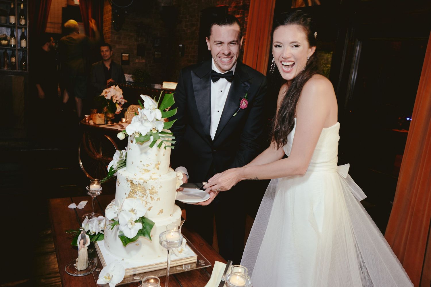 A couple in formal attire cuts into their elegant white tiered wedding cake adorned with fresh flowers.