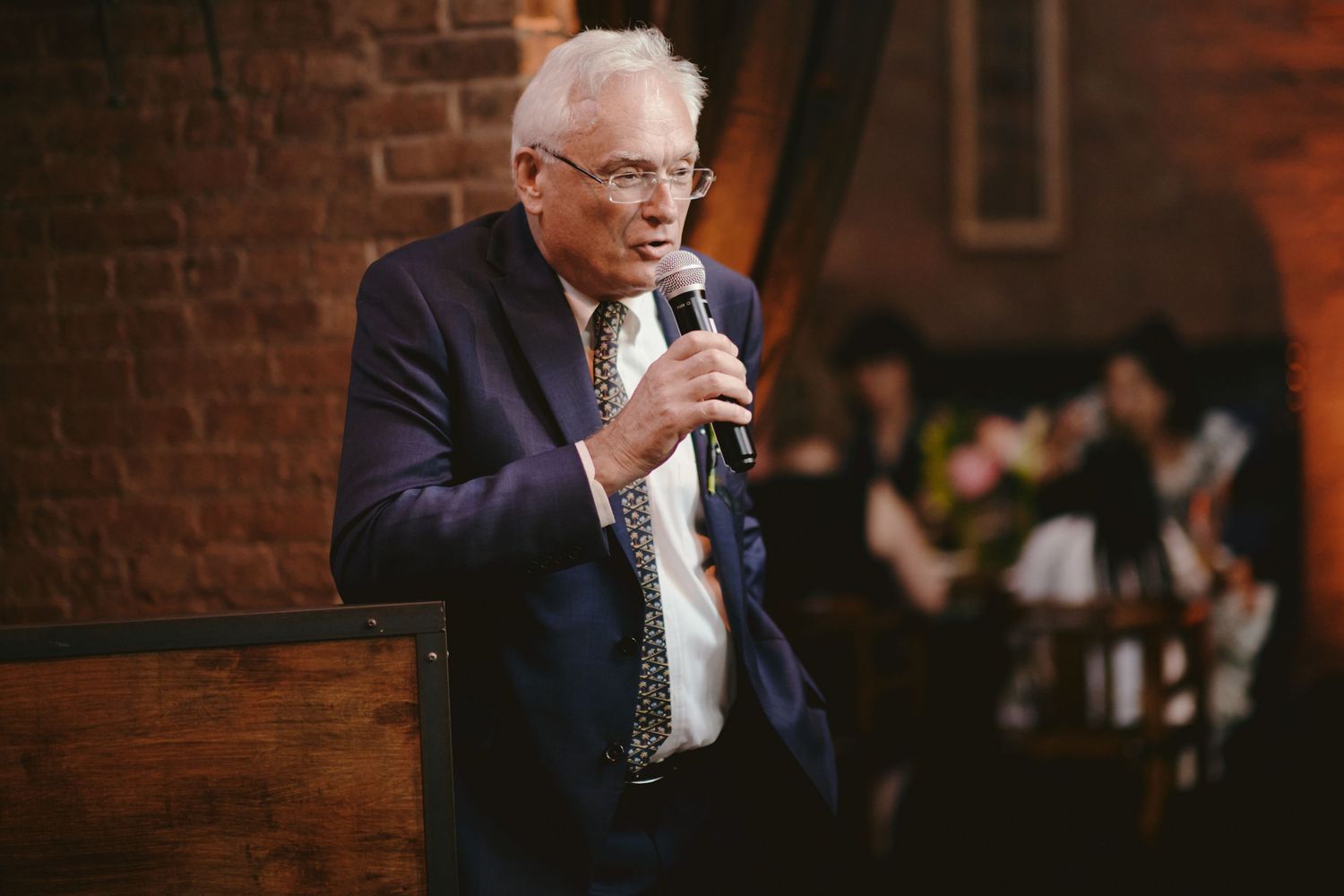 An elderly man in a suit speaks into a microphone at a brick-walled venue with people seated in the background.