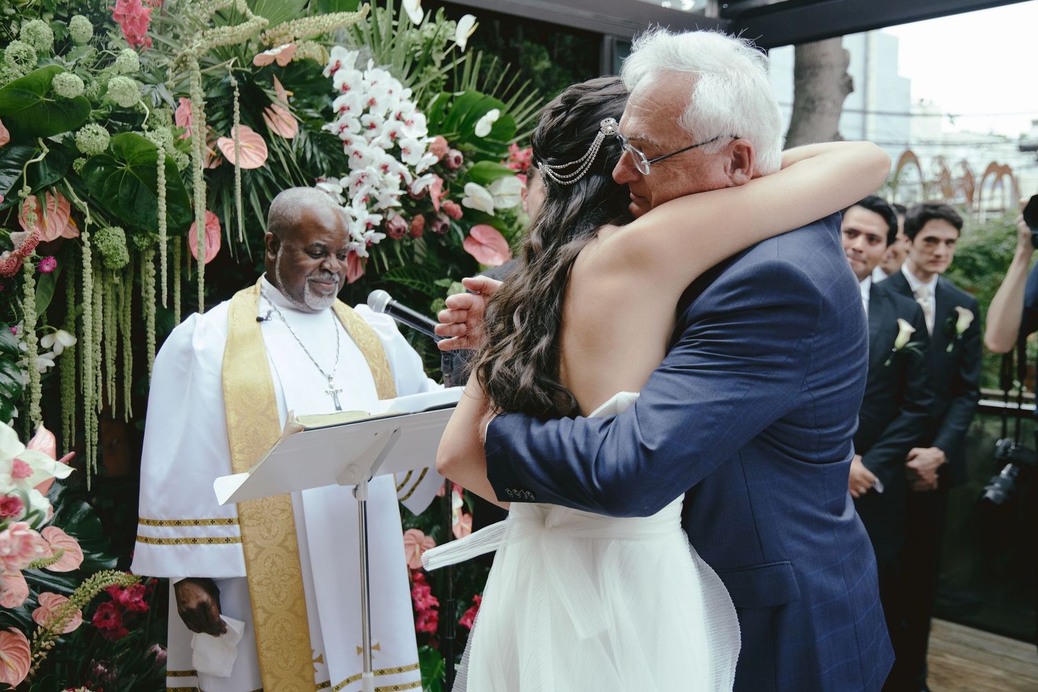 Two individuals embrace during a wedding ceremony officiated by a clergy member surrounded by floral decorations.