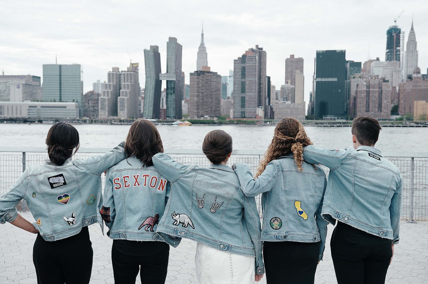 Five people wearing matching denim jackets face the New York City skyline along the waterfront.