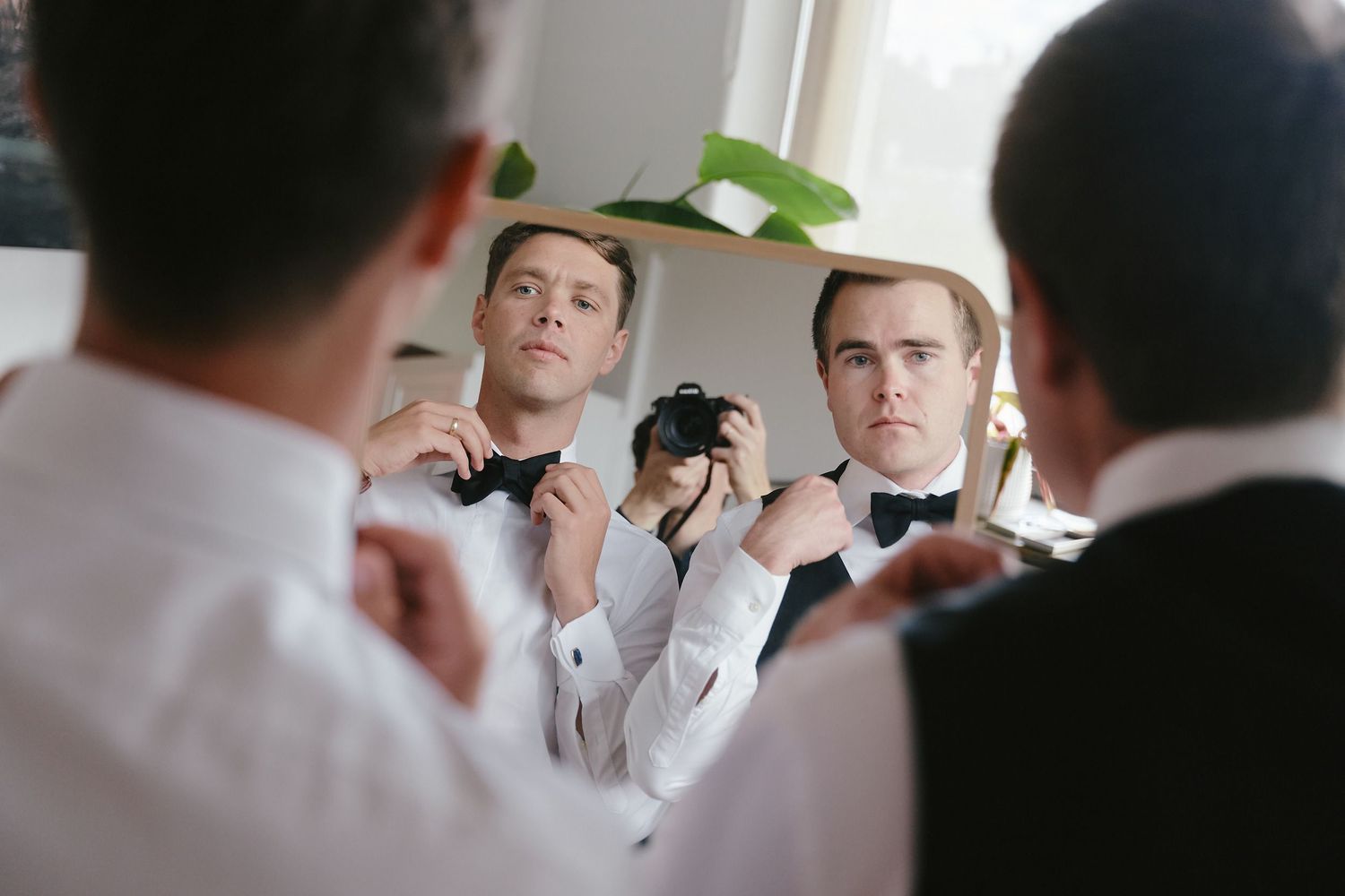 Groomsmen adjusting their bow ties in a mirrored reflection during wedding preparation.