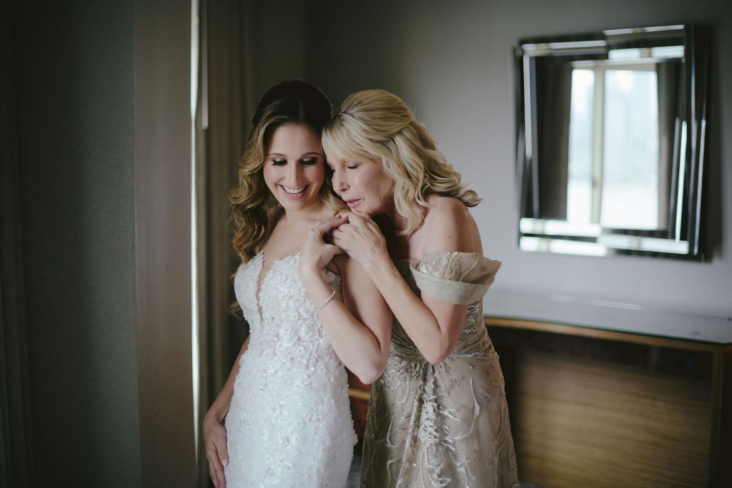 Bride in white lace gown gets help fastening dress from woman in beige gown in elegant hotel room.