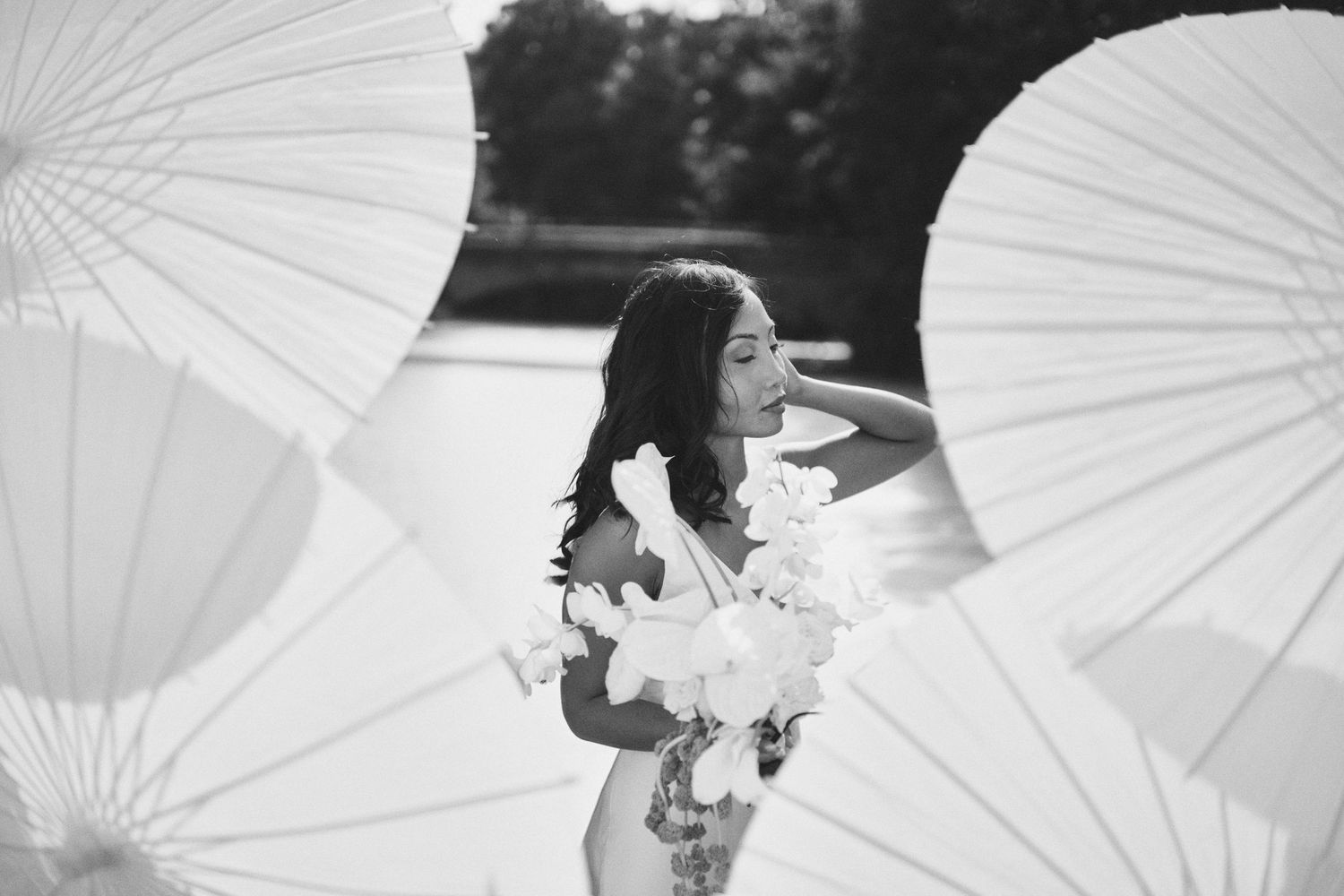 Black and white photo of a bride holding flowers surrounded by white parasols near a serene waterfront.