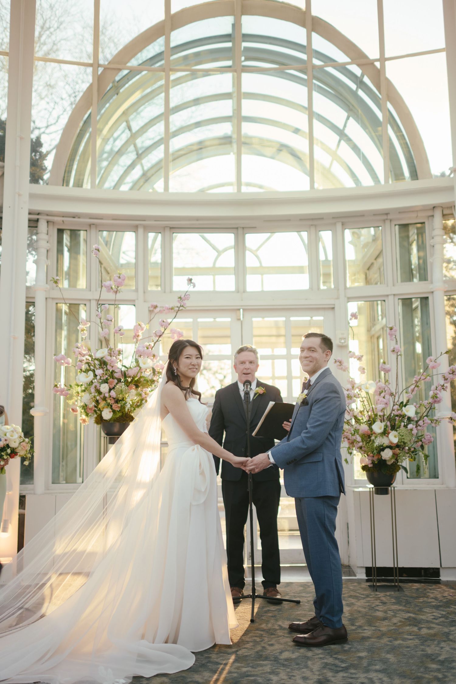 Bride and groom exchange vows in elegant glass conservatory chapel with cherry blossom floral arrangements.