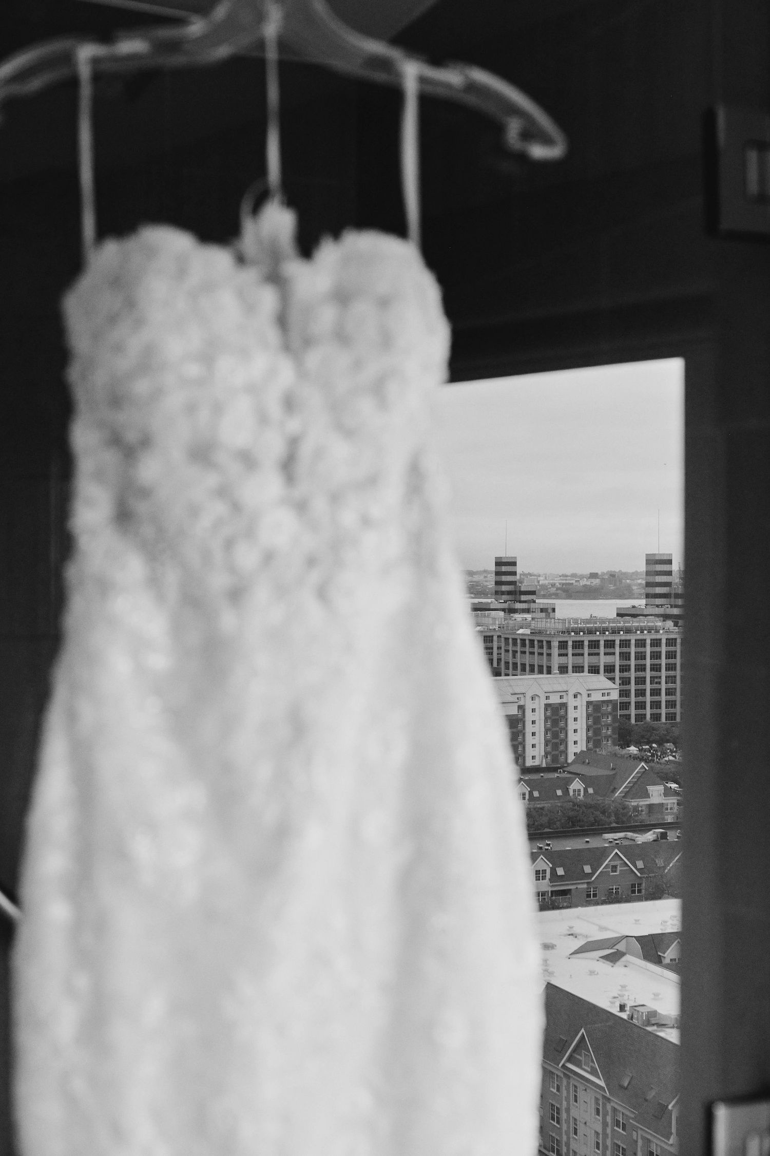 A white lace wedding dress hangs from a hanger near a window with a city view in black and white.