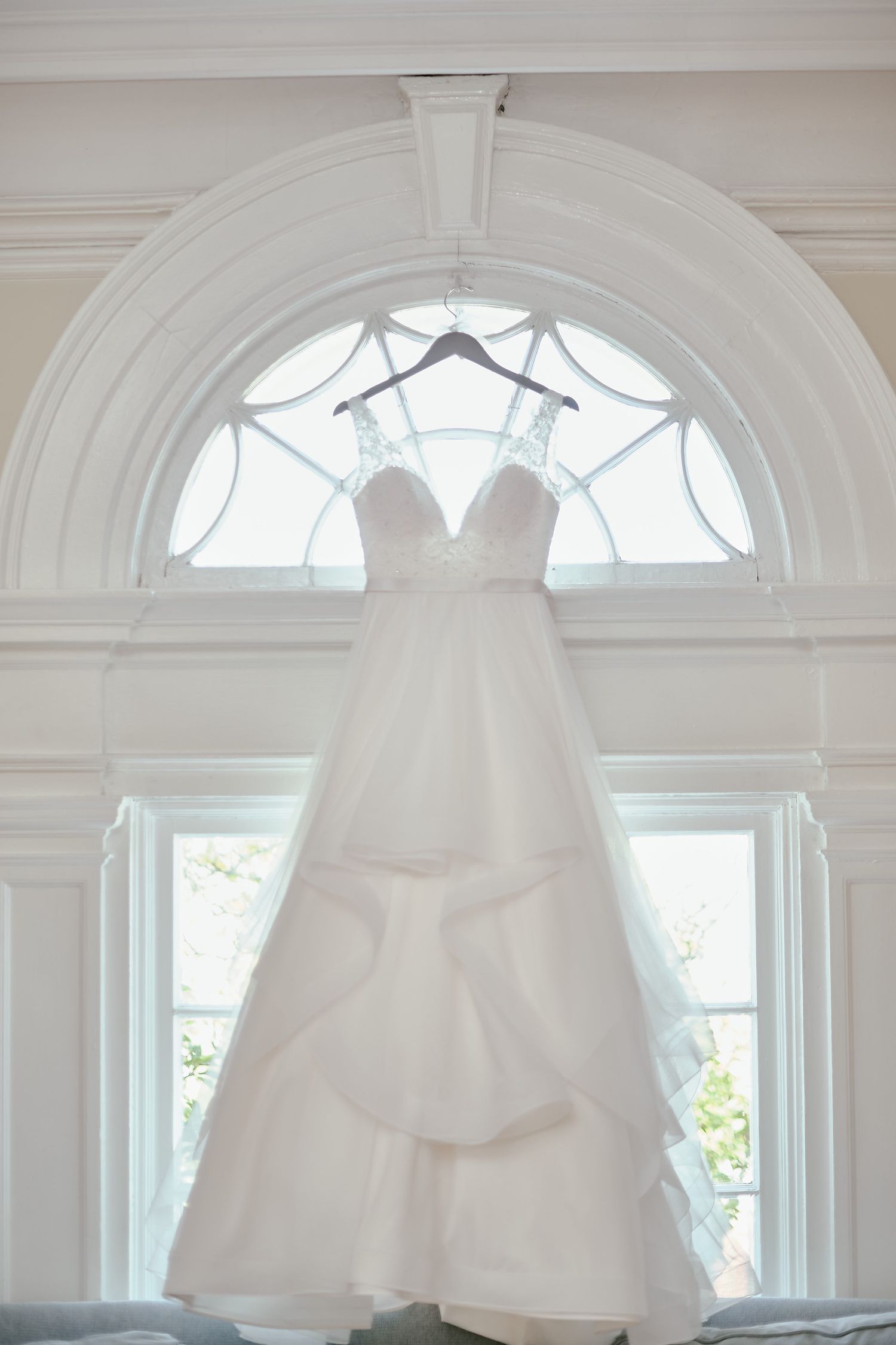 White wedding dress hanging elegantly in front of an arched window with decorative trim and natural light streaming through.