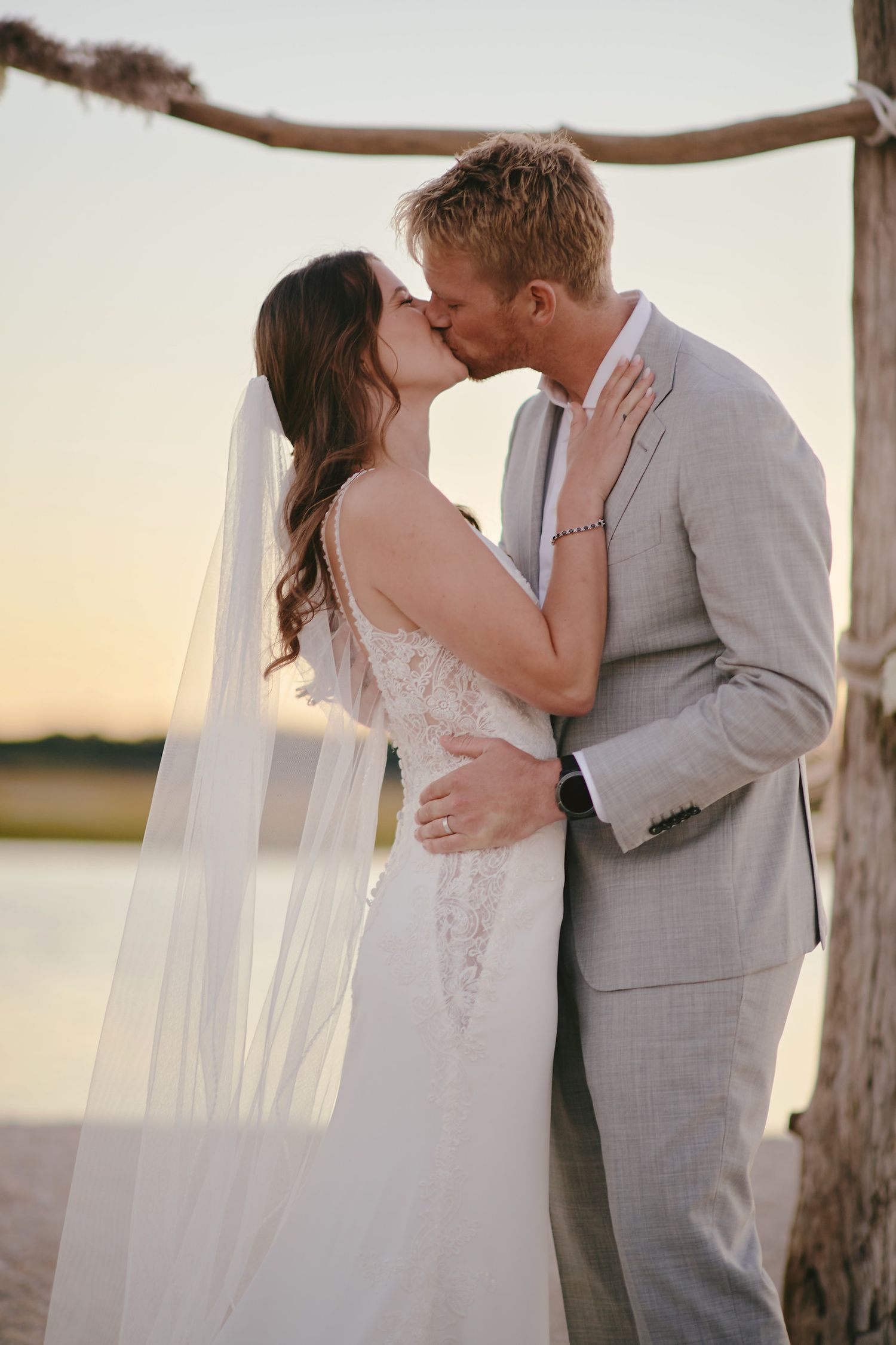 A romantic sunset wedding photo shows a couple sharing a kiss under a rustic wooden arch by a waterfront.
