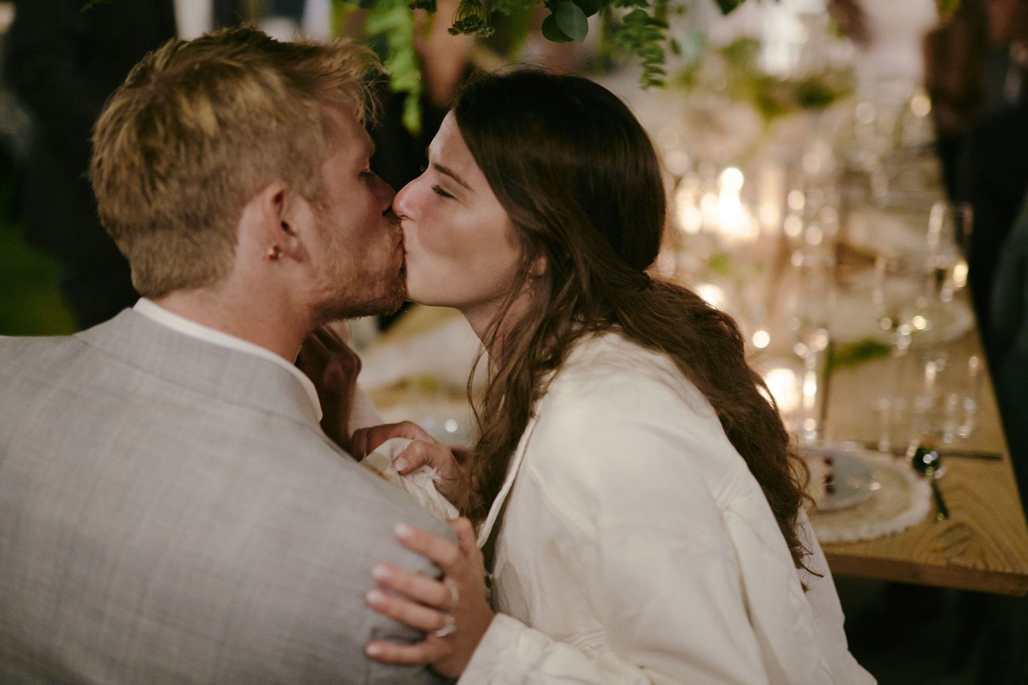 A romantic embrace between a couple in a soft lit restaurant setting with glowing candles and table settings in background.