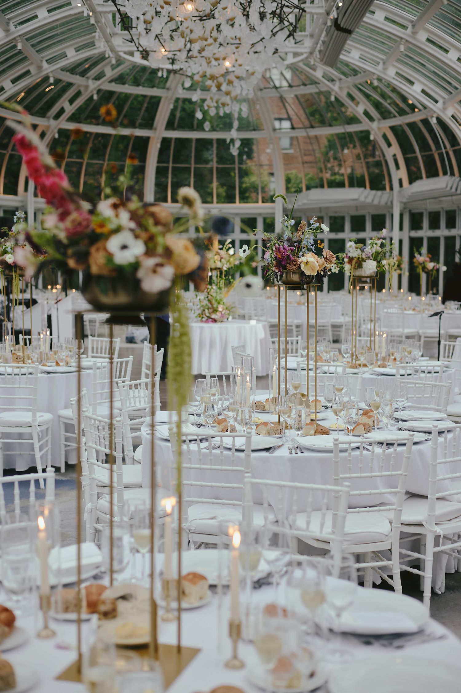 Elegant wedding reception setup inside a glass greenhouse with tall gold centerpieces and white chiavari chairs.