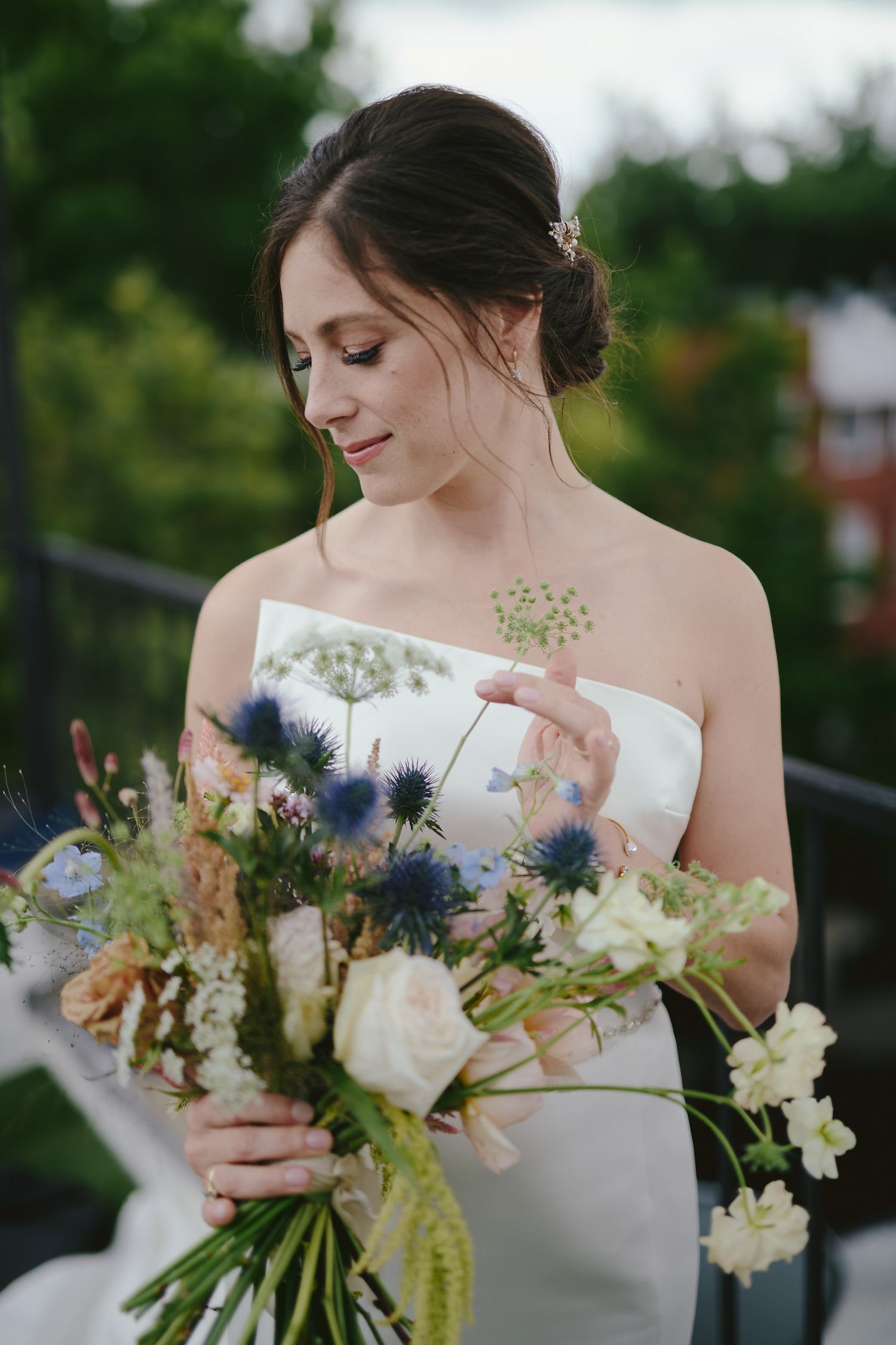 Bride admiring her elegant bouquet featuring blue thistles, white roses and delicate greenery against an outdoor backdrop.
