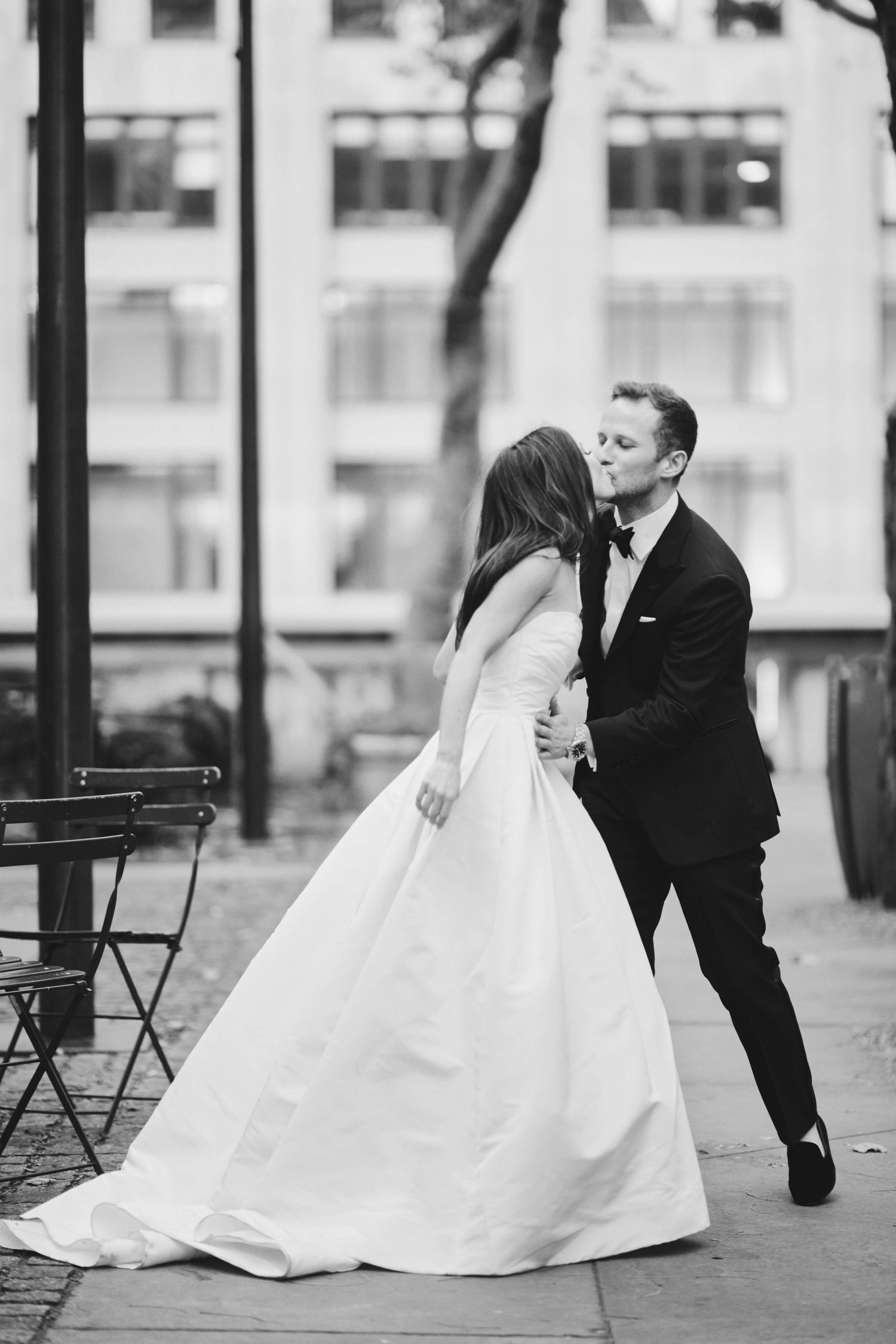 A romantic black and white wedding photo of a couple dancing in an outdoor urban setting with city buildings in background.