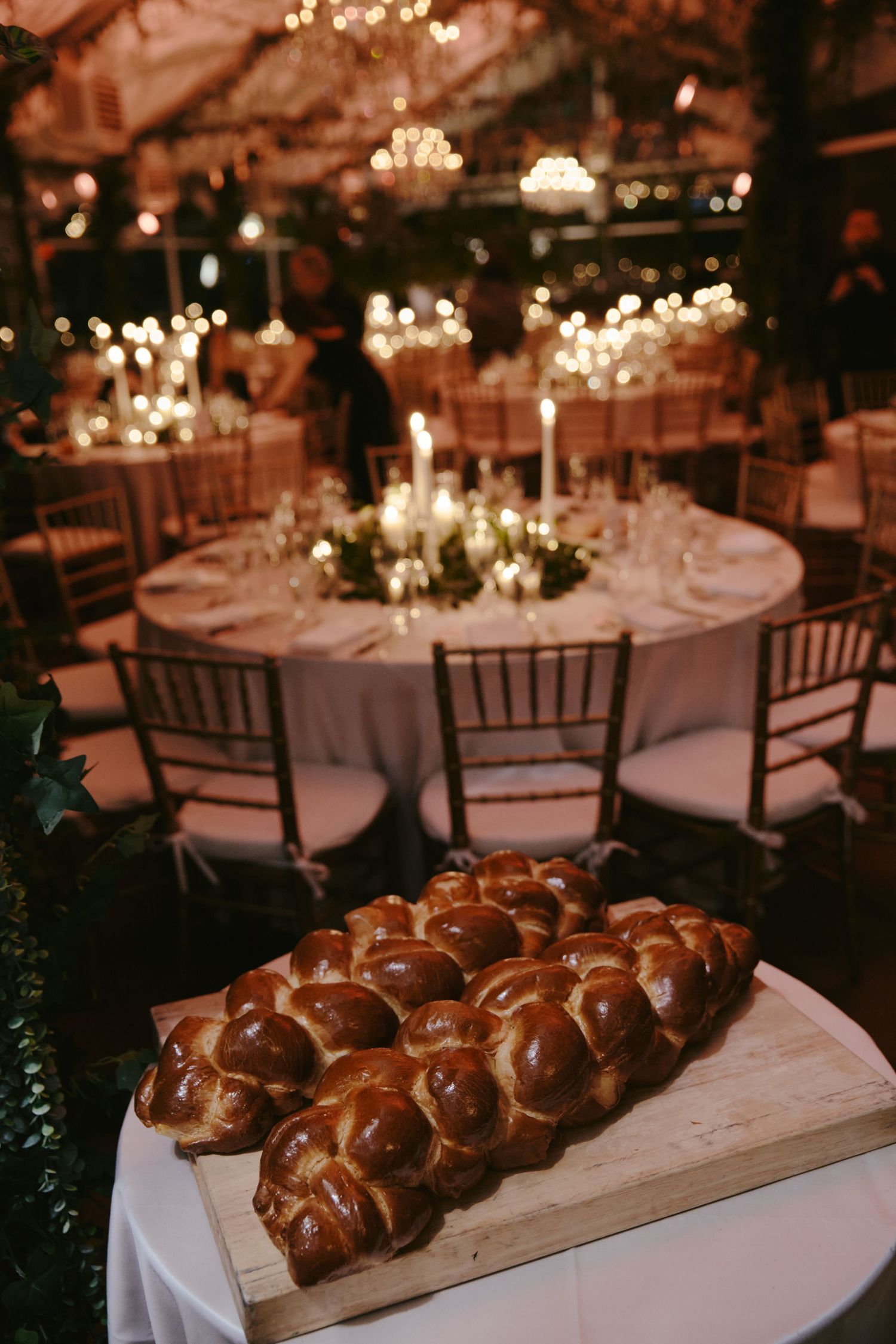 Fresh challah bread loaf displayed on wooden board at elegant wedding reception with round tables and candlelight decor.