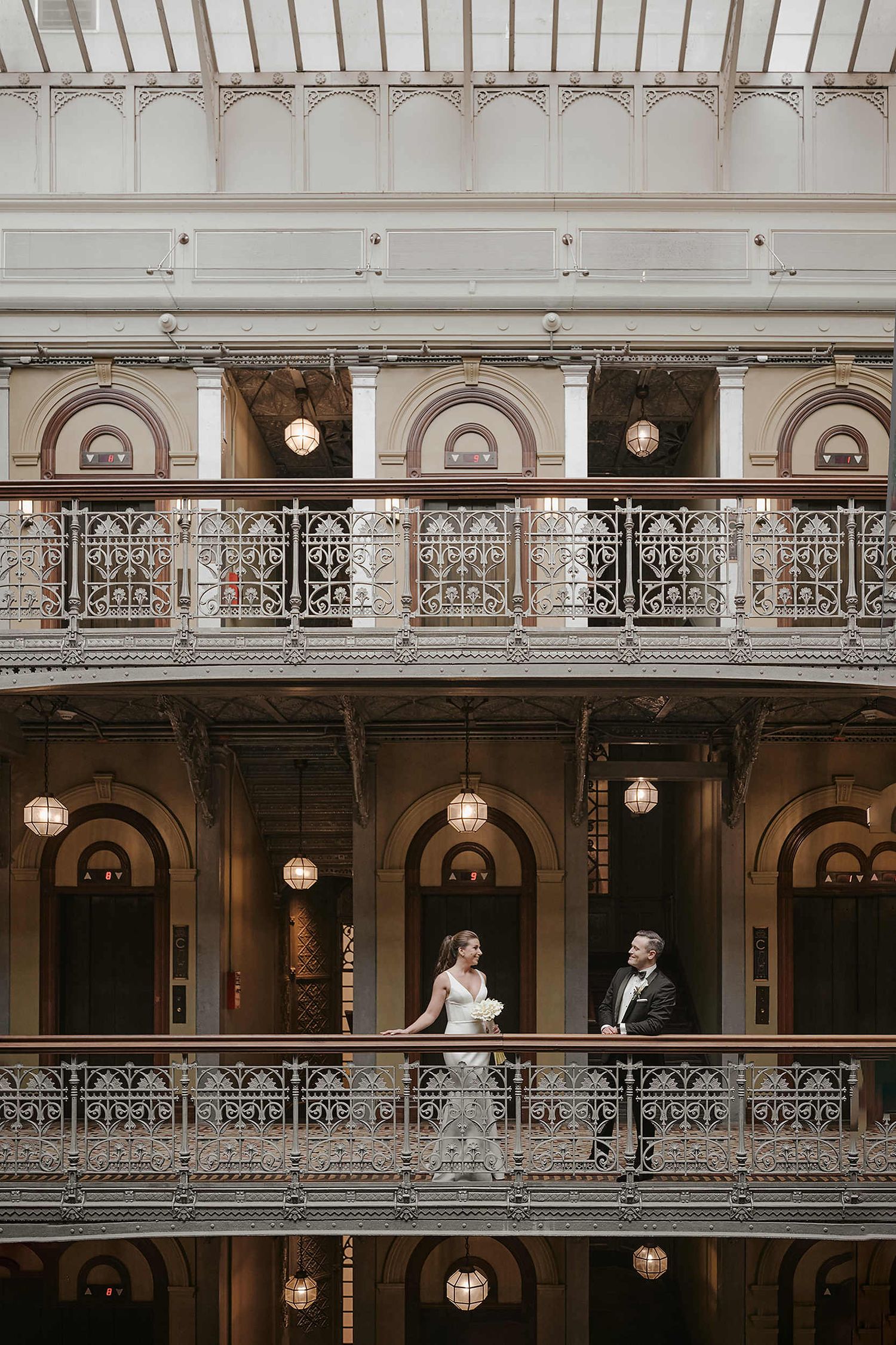 A couple poses on an ornate Victorian-style balcony with decorative iron railings and arched architecture lit by vintage lamps.