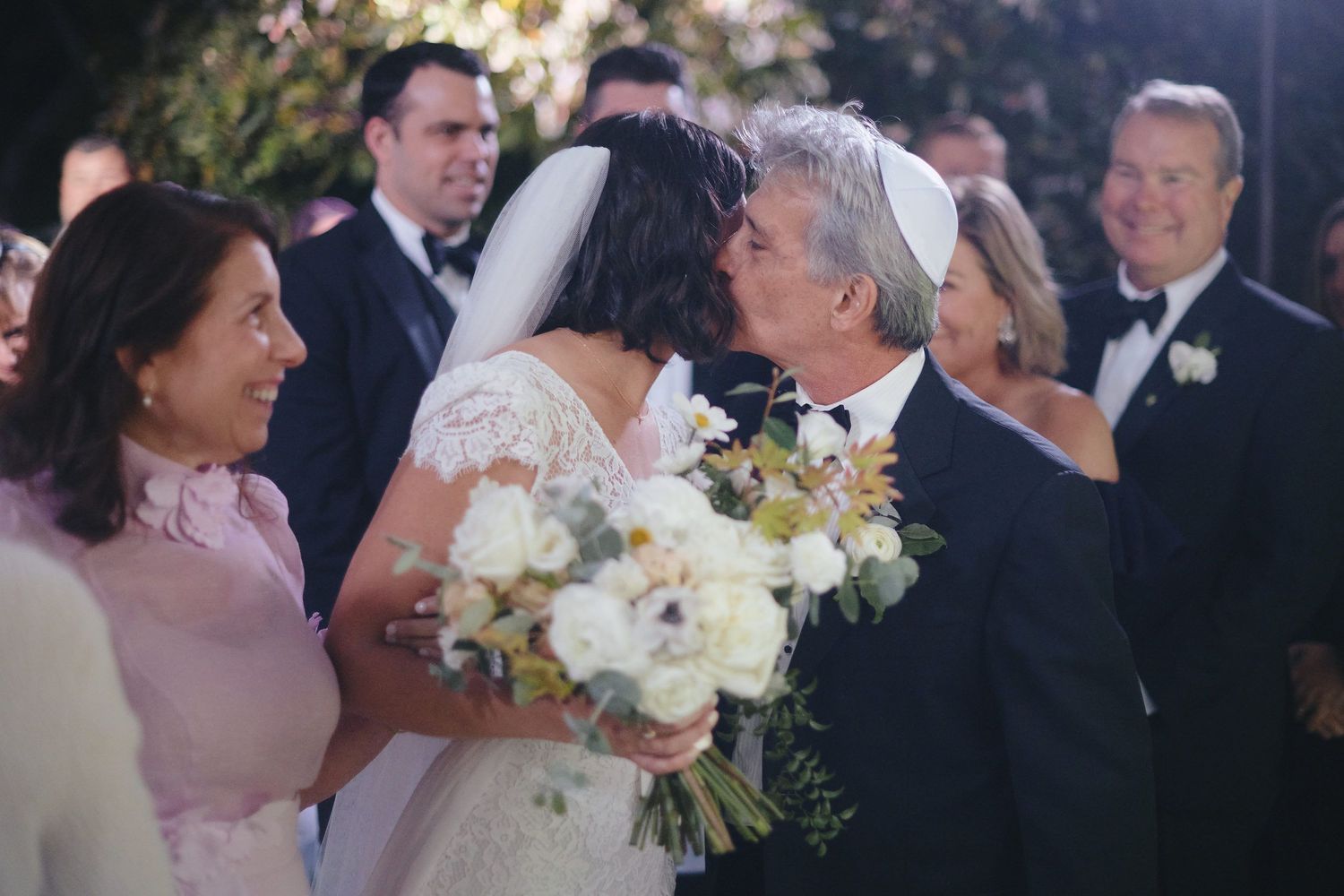A couple sharing a tender moment at their wedding celebration with white floral bouquet and guests in the background.