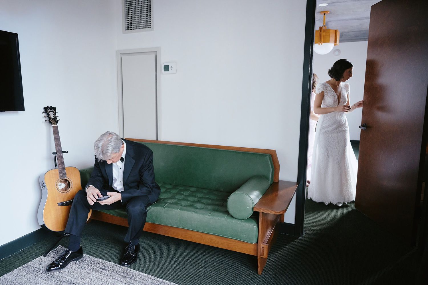 Two people wait separately in a room with a green couch and an acoustic guitar leaning against the wall.