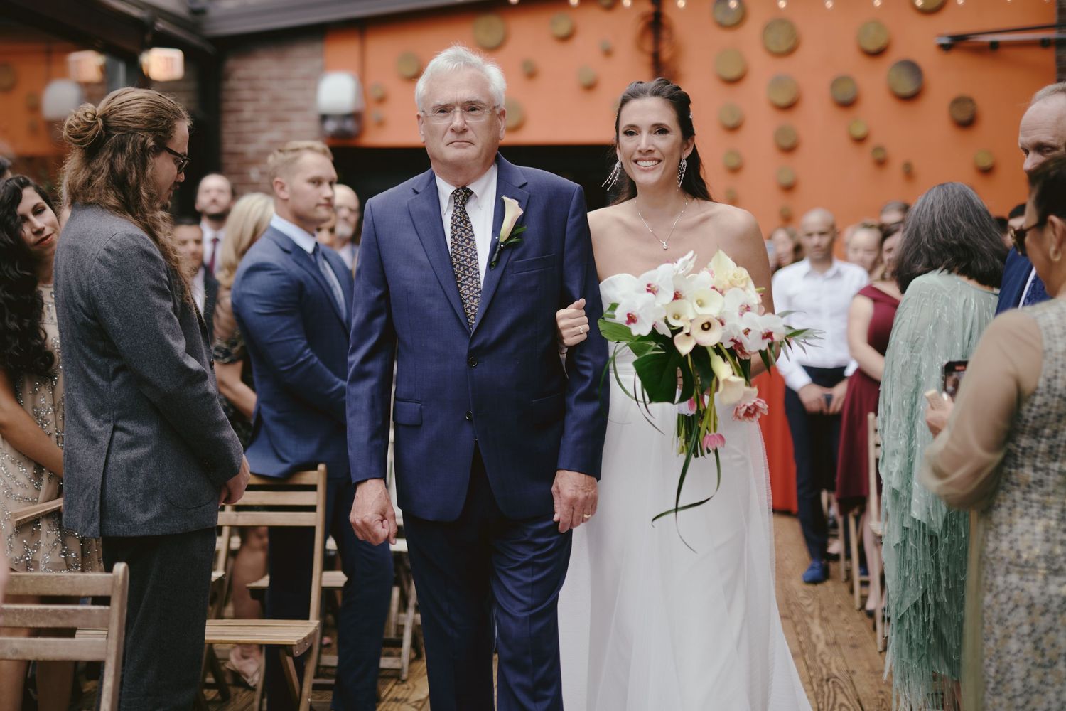 A wedding ceremony processional shows a couple walking down the aisle while guests look on from wooden benches.