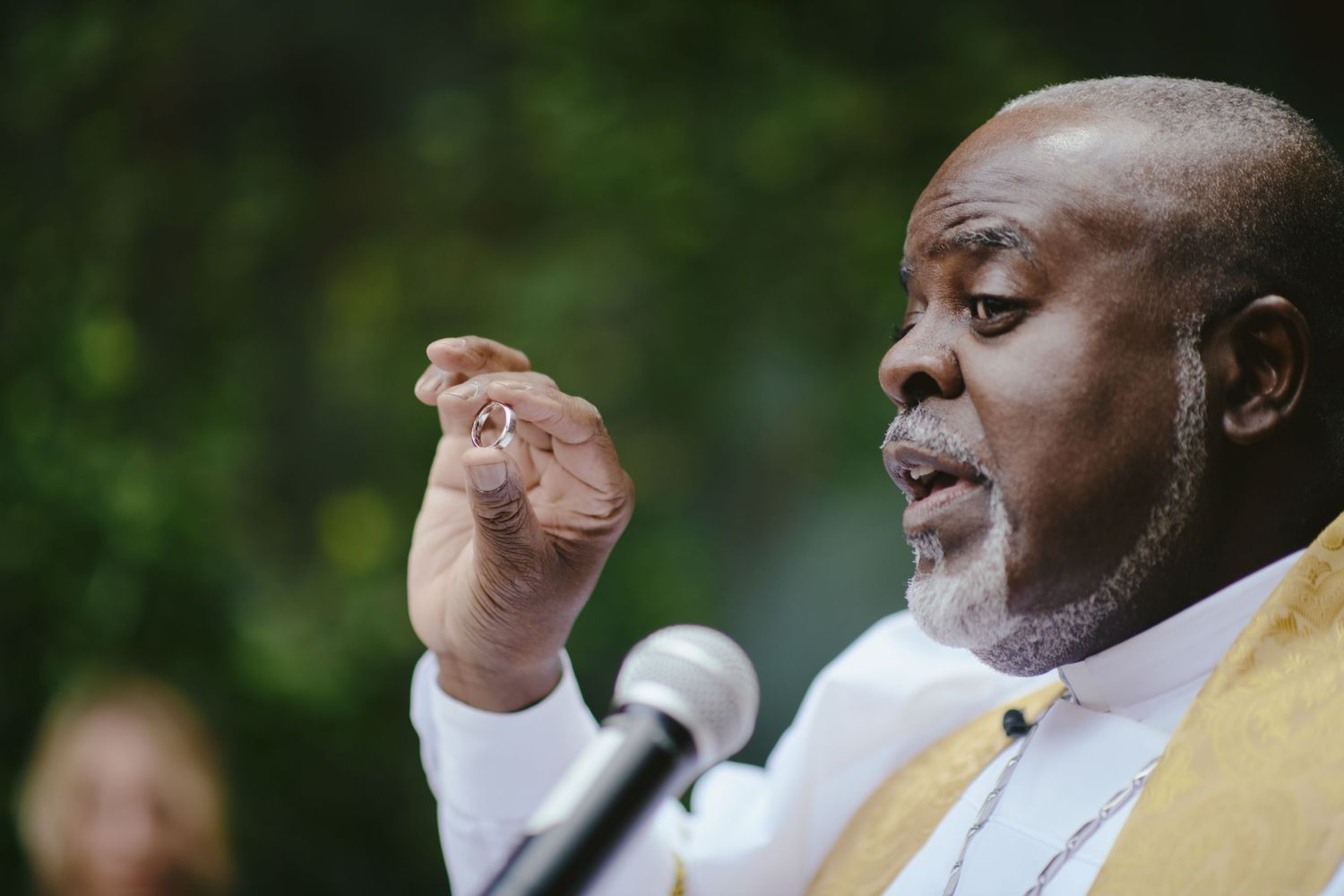 A speaker in a yellow vest gestures while speaking into a microphone against a blurred green background.