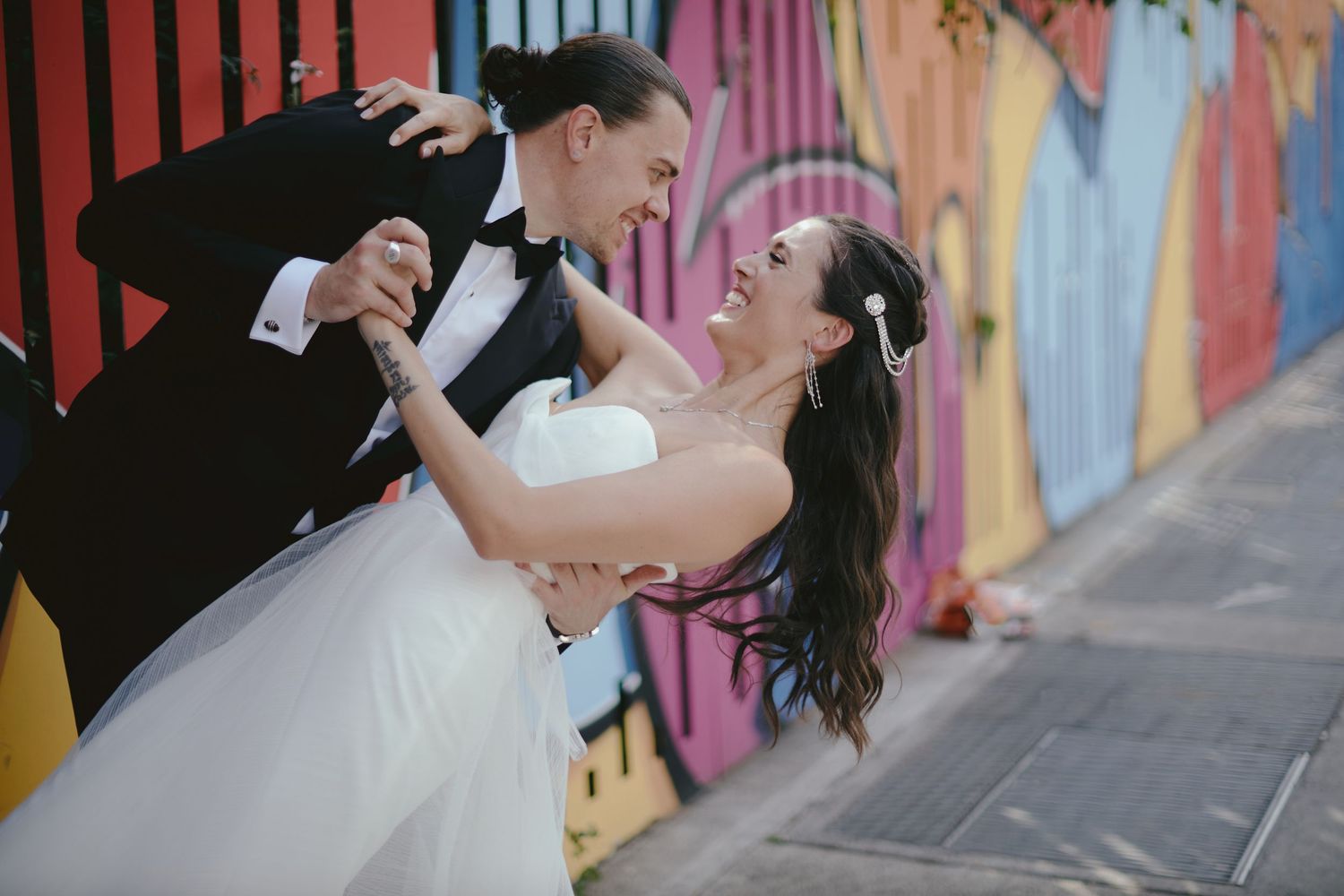 A happy couple in wedding attire dance and dip in front of a colorful street art mural wall.