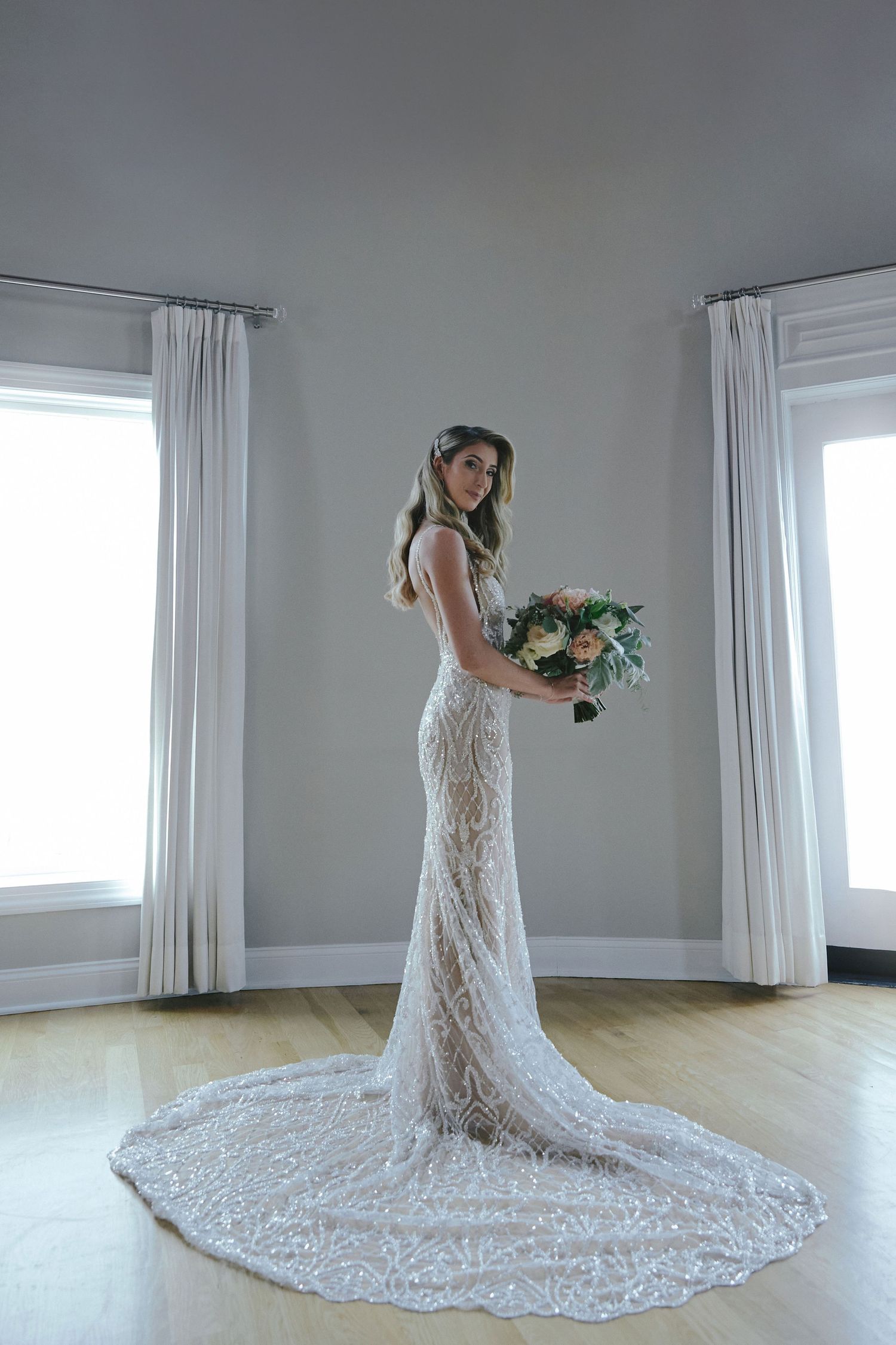 A bride in an elegant lace wedding gown with long train stands between two windows holding a floral bouquet.