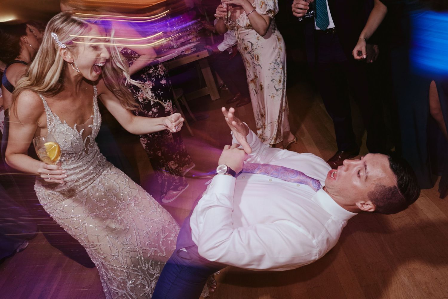Two people dance energetically and playfully during a reception under purple mood lighting on a dance floor.