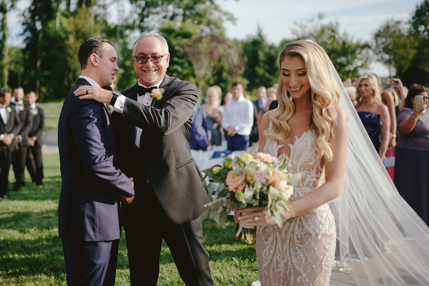 Outdoor wedding ceremony takes place on a sunny day with guests gathered on a green lawn.