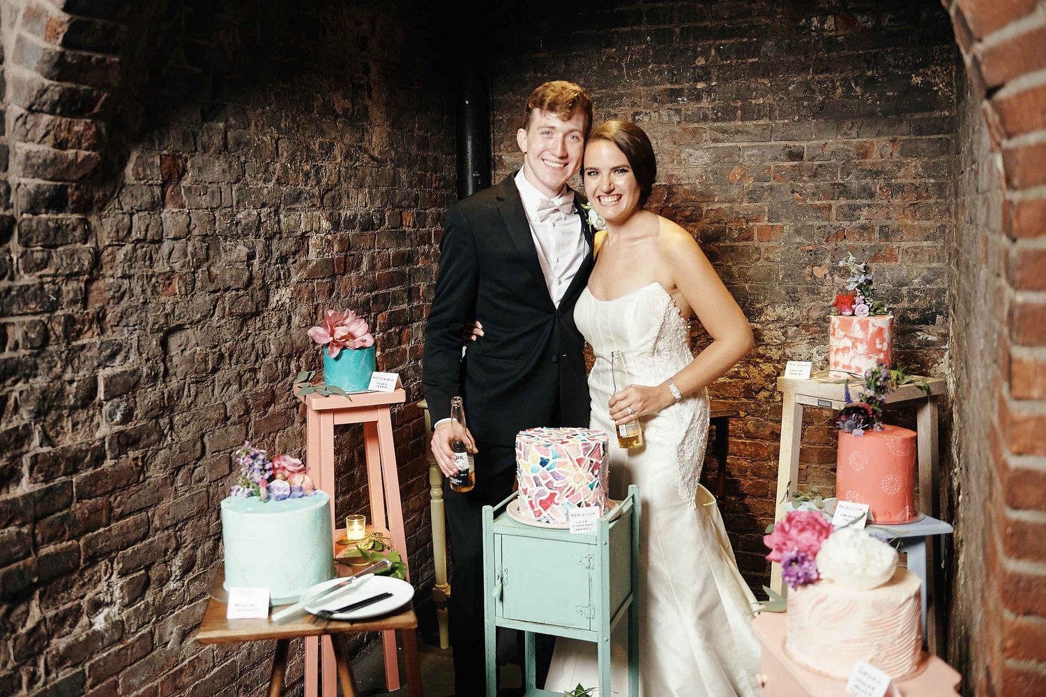 Vintage wedding scene with couple standing against brick wall surrounded by pastel decorative cakes on wooden stands.