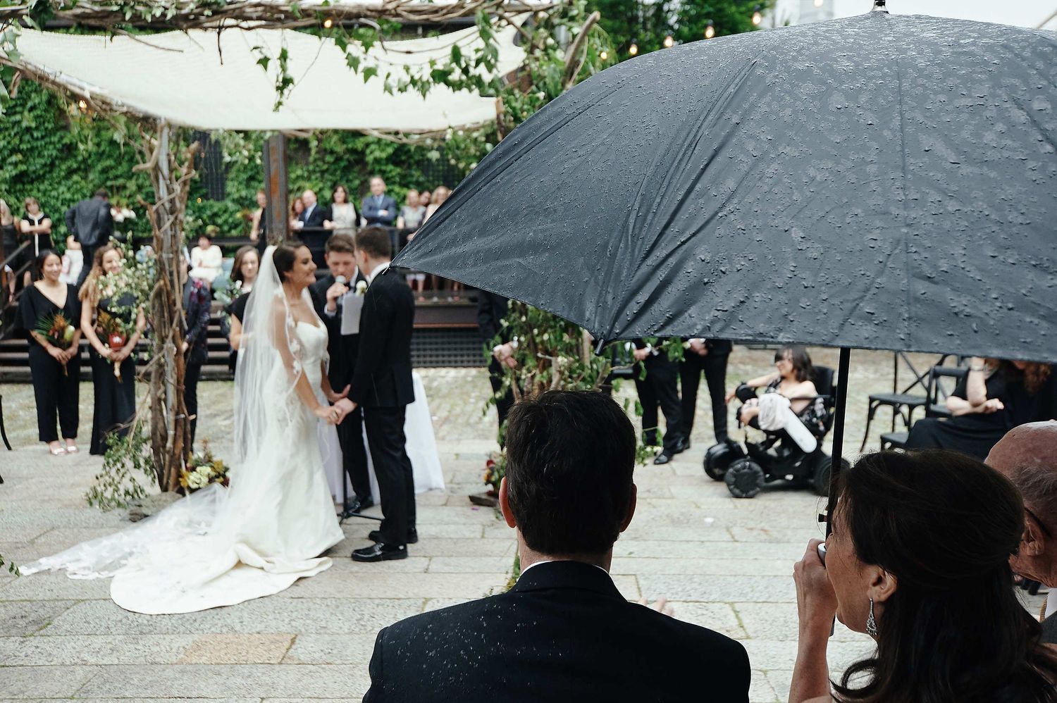 Wedding ceremony taking place under a black umbrella with guests watching in an outdoor garden setting.