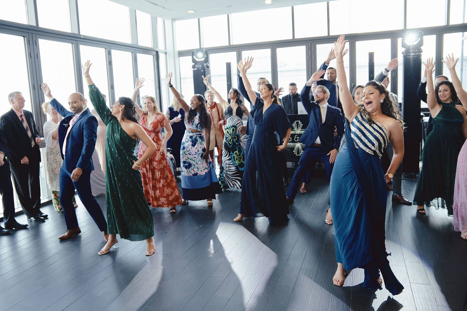 A group of wedding guests dancing with their arms raised in celebration in a bright room with floor-to-ceiling windows.