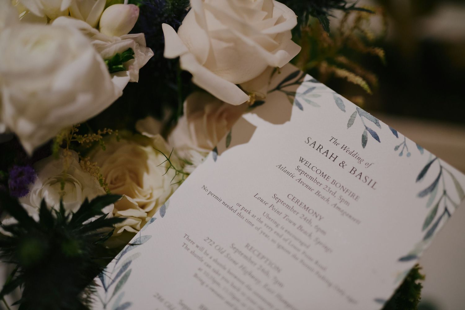 A wedding menu card with elegant script text lies beside white roses and florals in a romantic setting.