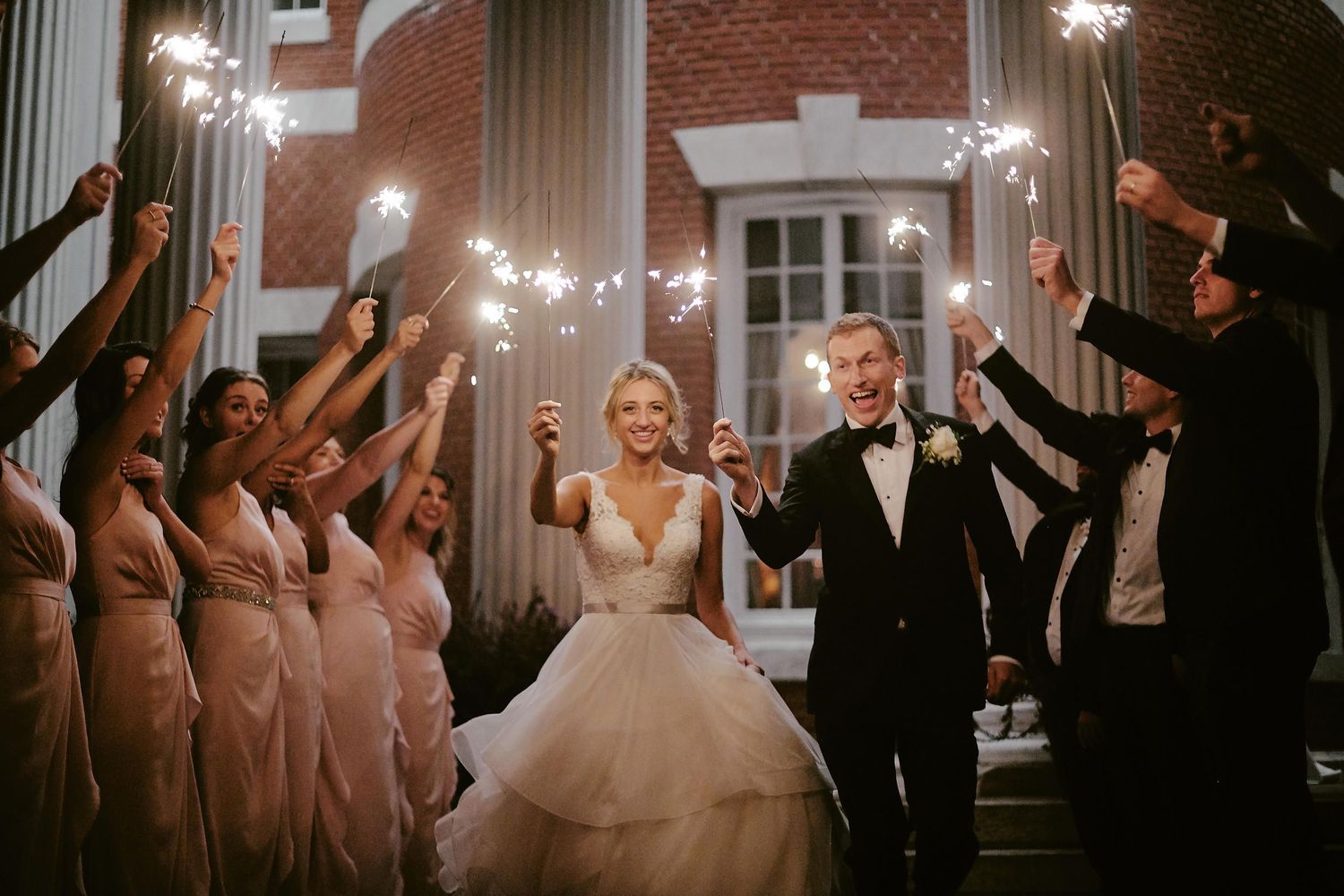Wedding party holds sparklers during evening celebration as couple walks through at elegant brick venue entrance.