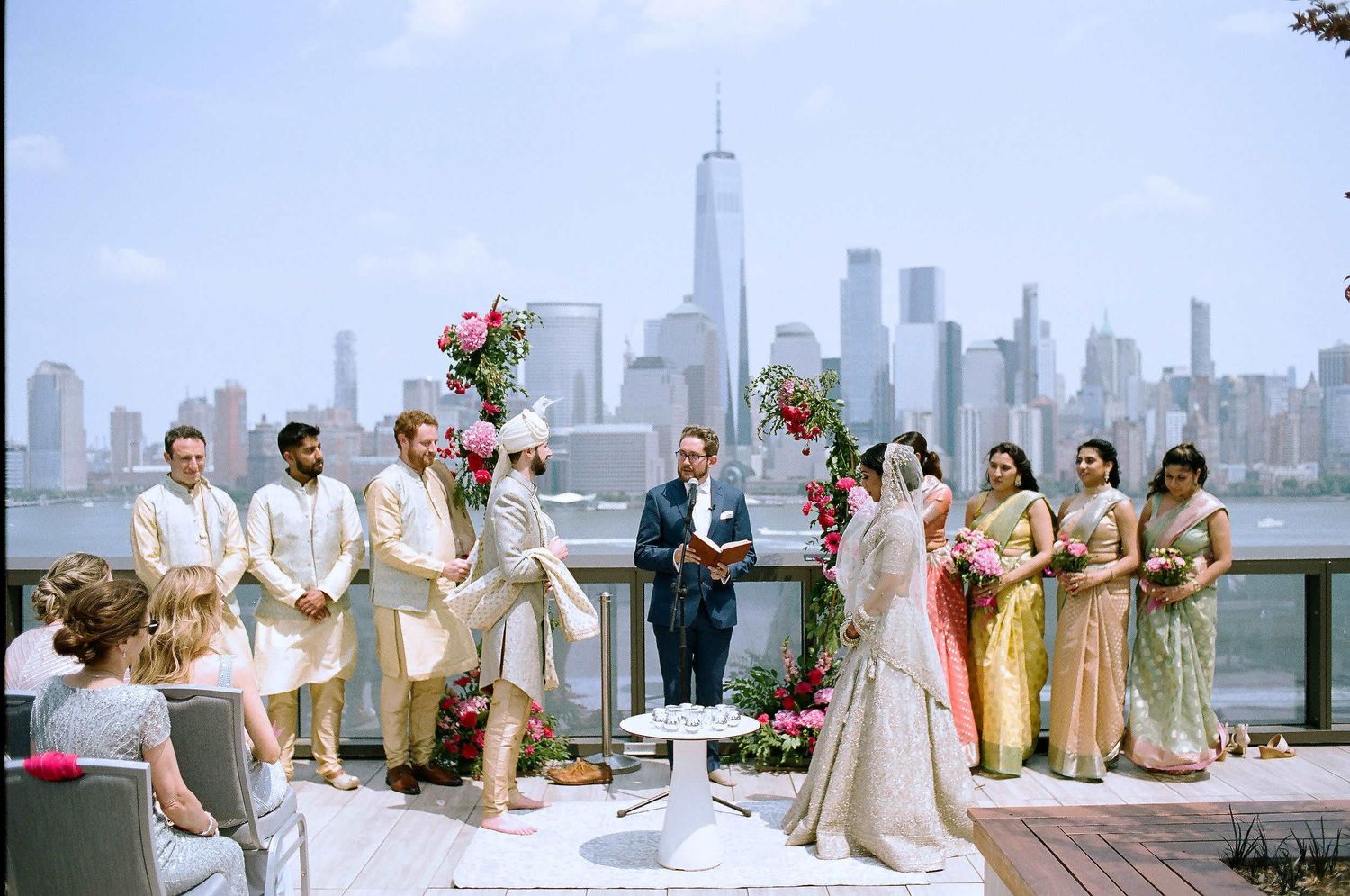 Wedding ceremony at waterfront with New York City skyline and One World Trade Center in background on sunny day.
