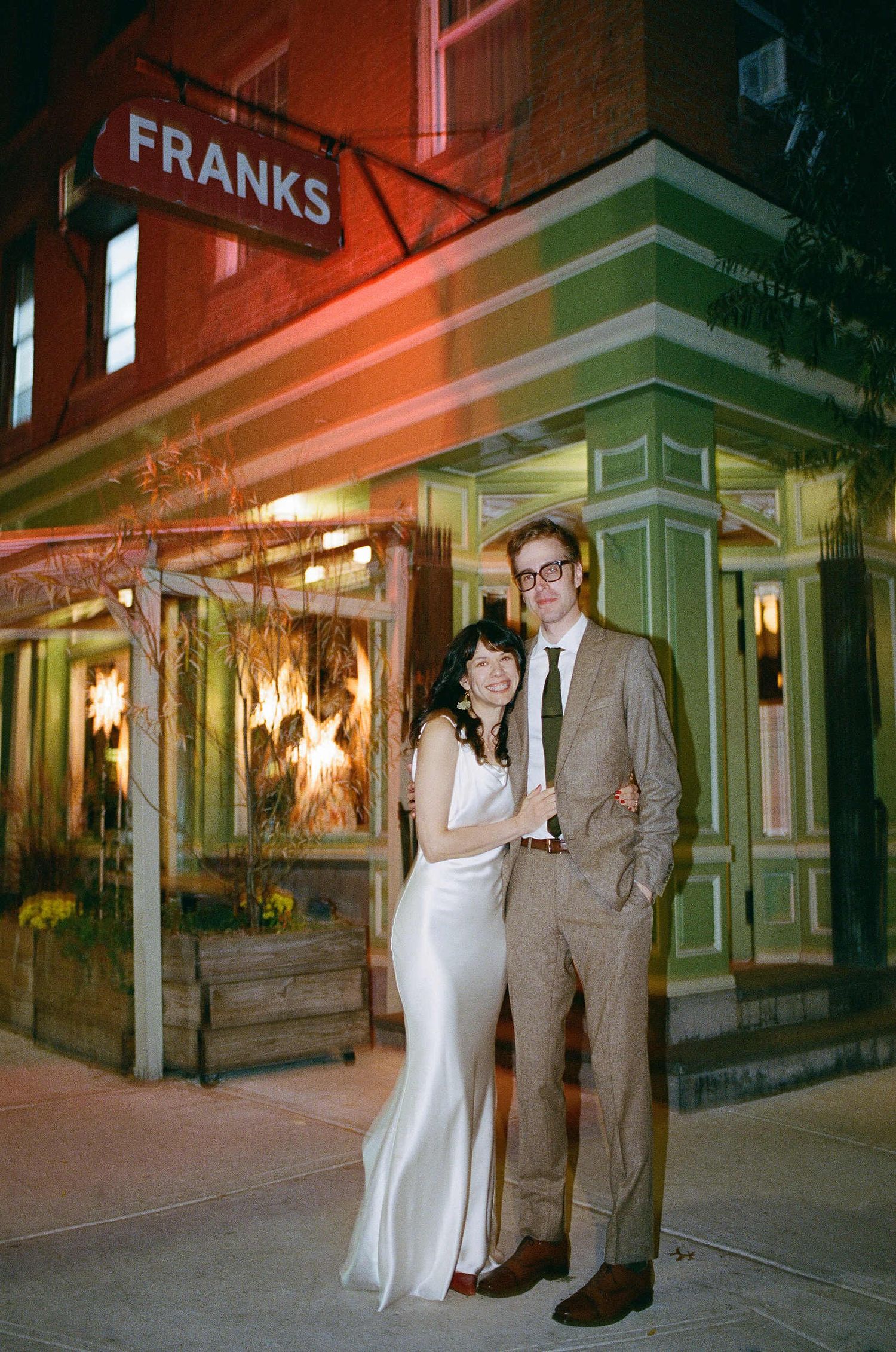 A couple poses outside Franks restaurant at night with glowing lights and vintage green storefront details.