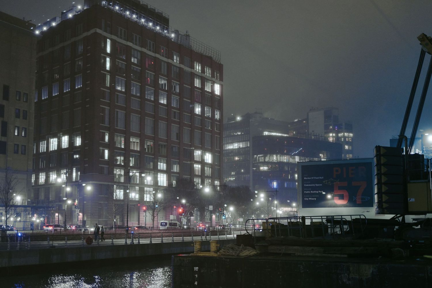 Moody night cityscape with illuminated brick building and industrial crane along misty waterfront in dark urban setting.