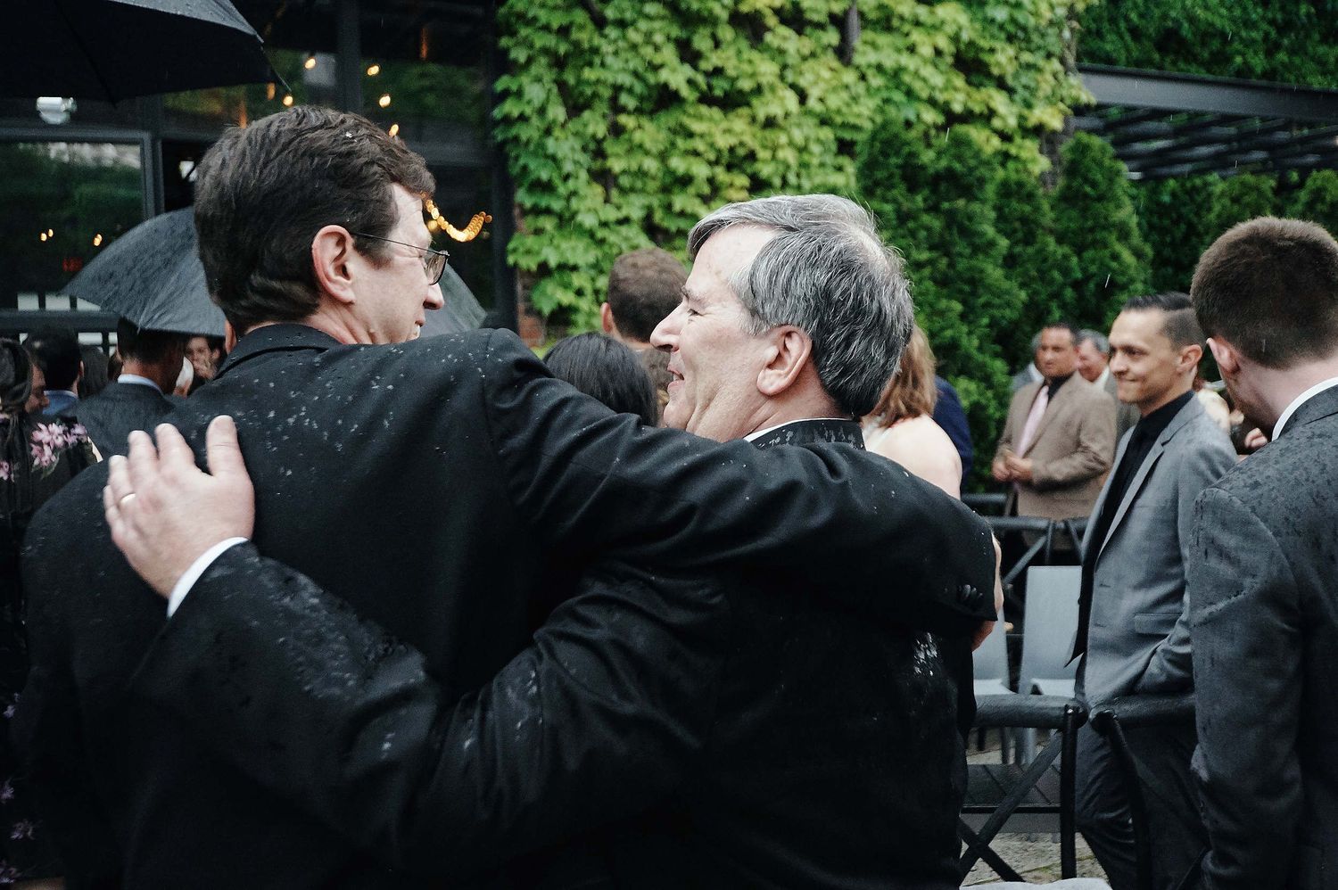 Two men share an emotional embrace at an outdoor gathering surrounded by other formally dressed guests.