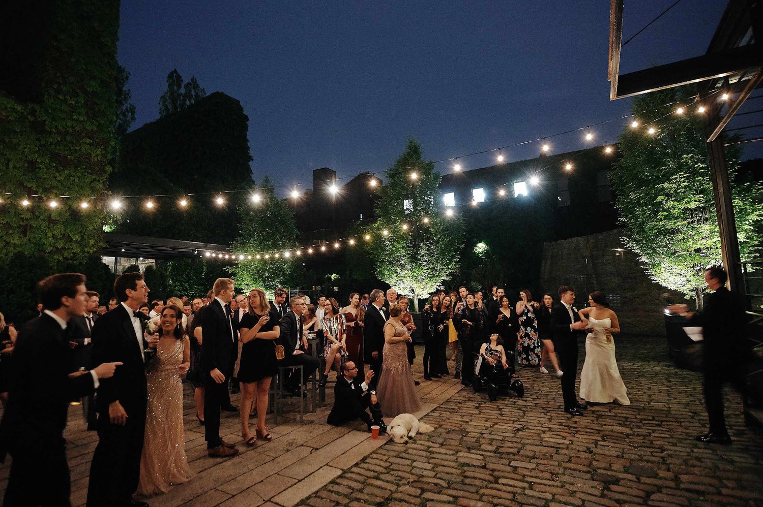 A nighttime outdoor wedding reception with string lights illuminating guests dancing on a cobblestone patio.