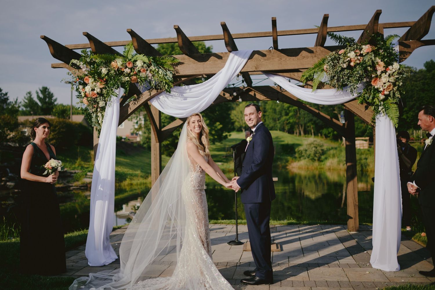 A outdoor wedding ceremony takes place under a wooden pergola decorated with white draping fabric and floral arrangements.