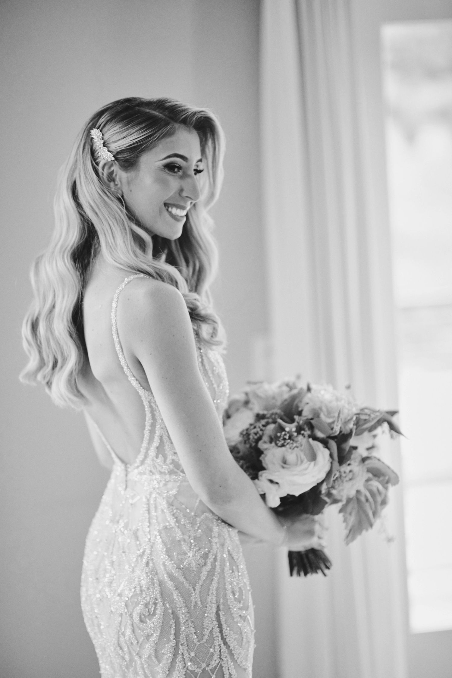 Black and white photo of a bride in a fitted lace dress holding a bouquet while smiling beside a window.
