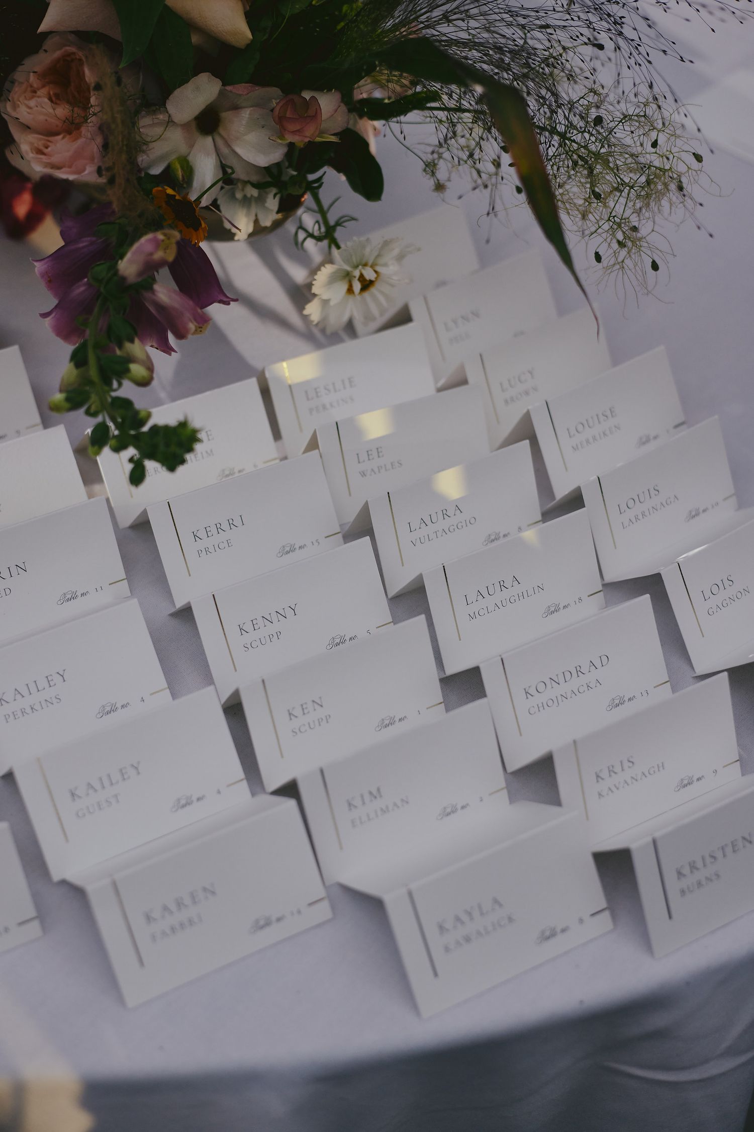 Wedding place cards arranged in neat rows on a white surface with delicate floral arrangements in the corner.