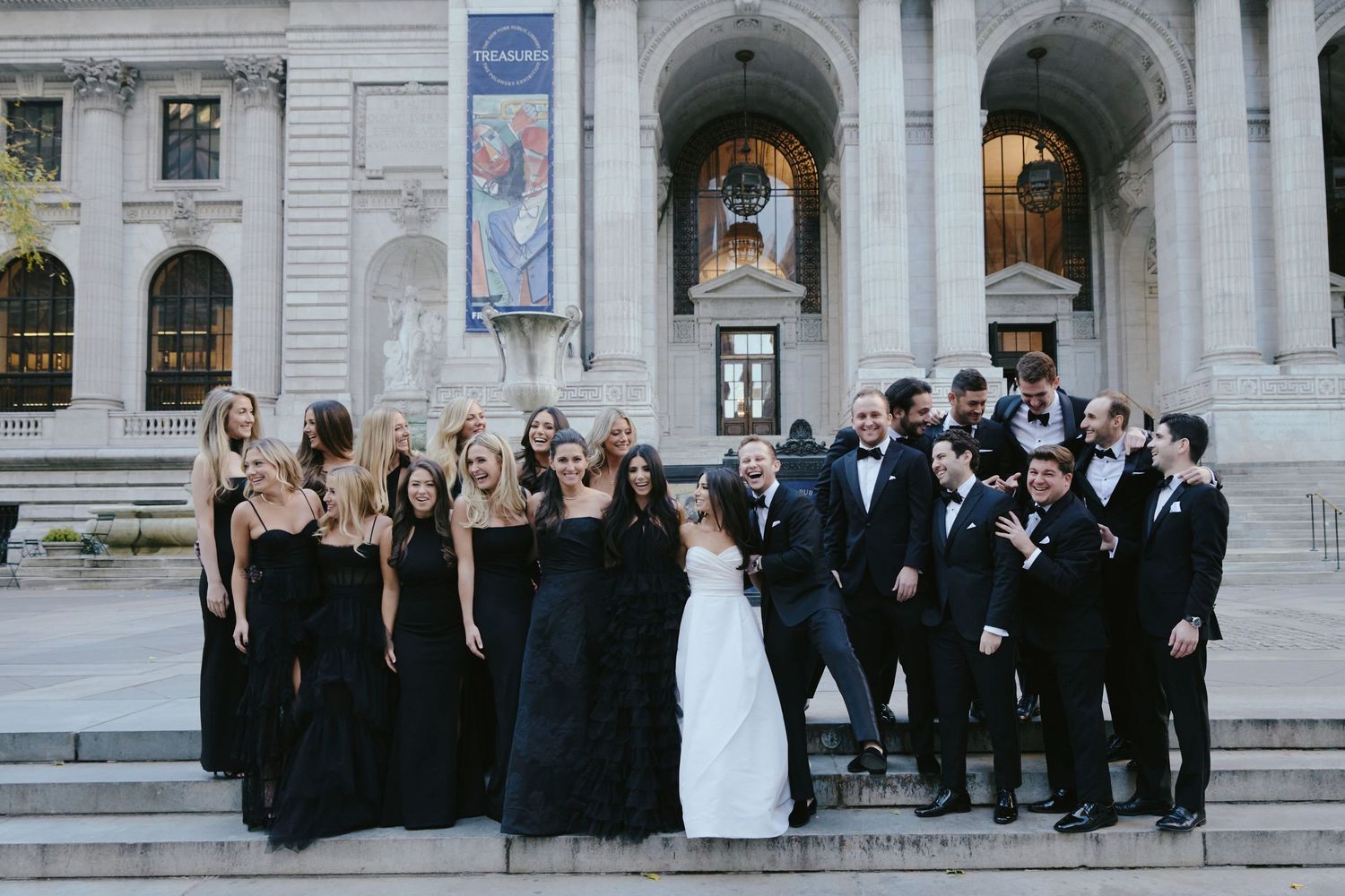 Wedding party in formal attire poses on grand stone steps of historic building with arched columns.