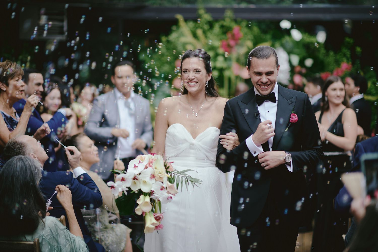 A couple walks down the aisle surrounded by guests throwing white confetti petals at their outdoor wedding celebration.