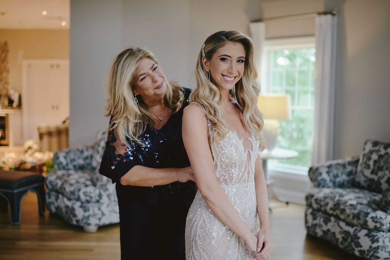 A tender moment captured as a woman helps adjust a fitted lace wedding gown in a sunlit room.